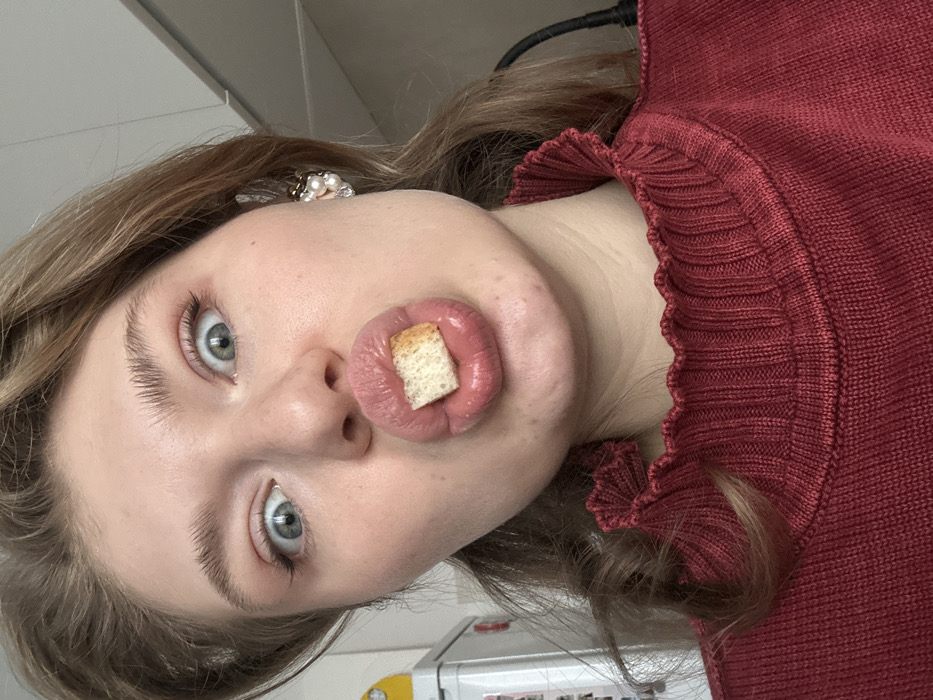 An indoor, close-up, head-and-shoulders portrait depicts a fair-skinned female, approximately 20-30 years old, with light brown hair, wide blue eyes, and light makeup, including defined eyeliner. Her lips are puckered into a "fish-face" pose, holding a small, rectangular piece of white bread or crouton. She is looking directly forward into the camera. She wears a dark red, high-necked, ribbed knit top with a ruffled collar. A decorative earring, appearing to be composed of small pearls and possibly other embellishments, is visible on her right ear. The background is out of focus, revealing a white appliance (possibly a refrigerator) to the left and a dark object or cable to the right, suggesting a domestic setting such as a kitchen. The scene is located in Kazan, Russia.