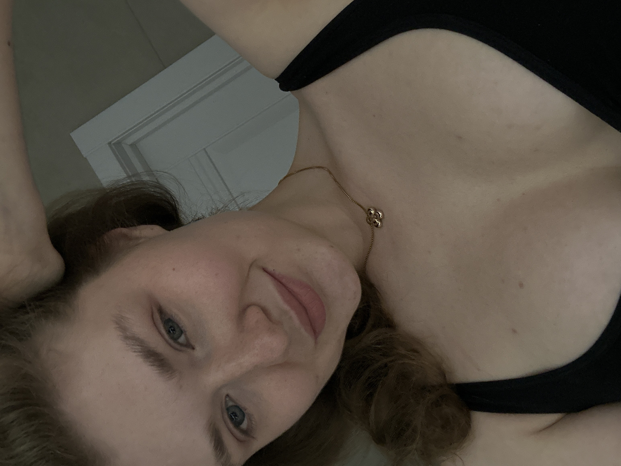A selfie photograph taken indoors featuring a young woman. She has fair skin, blue eyes, and light brown hair. She is wearing a black top and a gold necklace with a flower-shaped pendant. The background shows a wall and a door frame. There is no information in the image that indicates the location is Kazan, Russia.