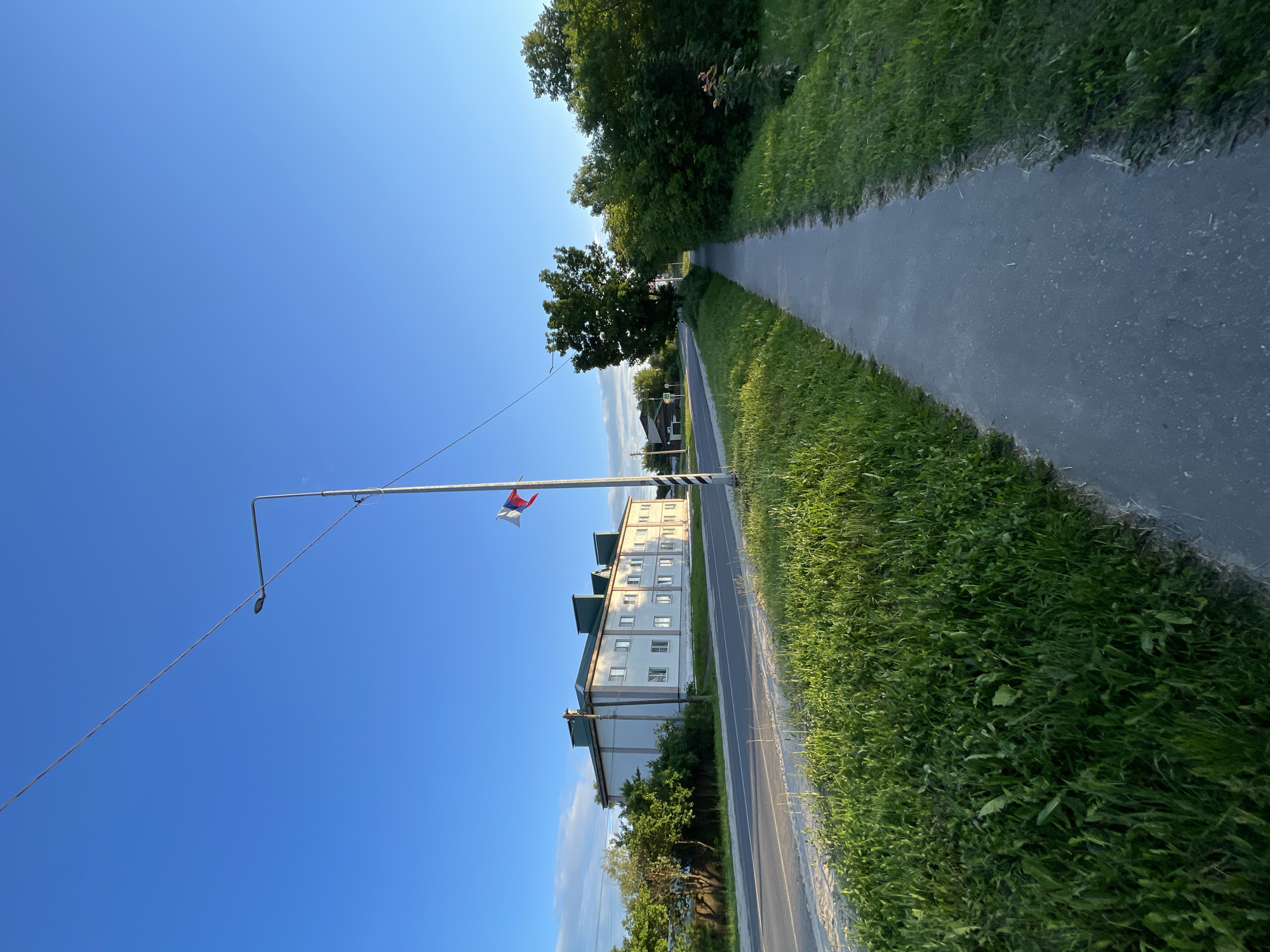 The image captures a street scene in Krasnye Baki, Russia, under a clear blue sky. A paved sidewalk runs along the right side of the frame, bordered by a strip of lush green grass. Across the street, a long, white, multi-story building with a green roof dominates the left side of the scene. A road separates the building from the sidewalk and the grassy verge. A lamppost stands on the verge near the sidewalk, with a small flag, appearing to be the Russian flag, attached to it. Trees and bushes line the right side of the sidewalk, and more greenery is visible behind the building on the left. The sun appears to be low in the sky, casting a warm light on the scene.