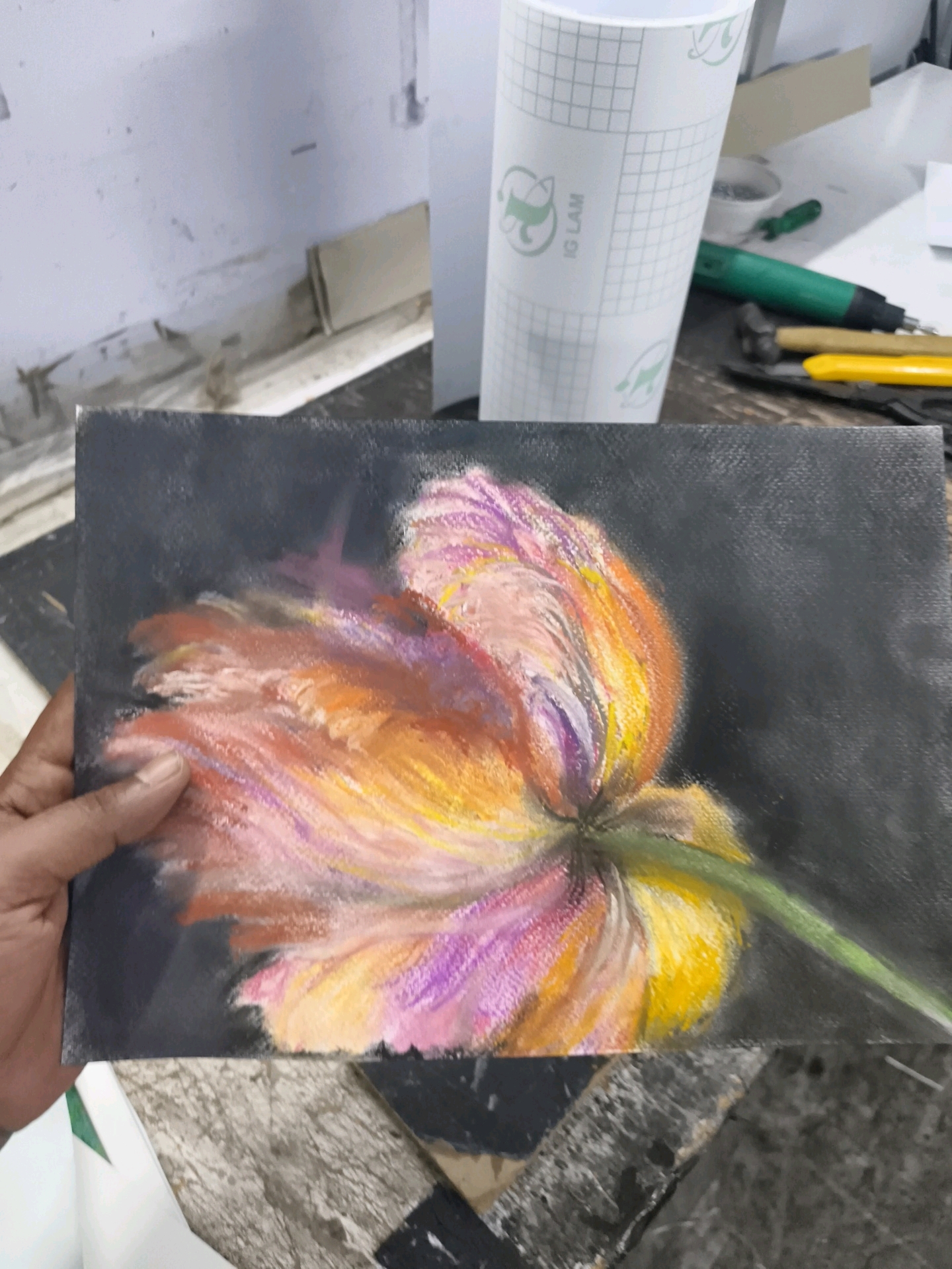 A pastel painting of a vibrant tulip is held by a person's left hand against a dark background. The tulip features a blend of orange, pink, and yellow petals with touches of purple and green. The painting is on a canvas and is placed on a surface that appears to be a wooden table, possibly in a workshop or art studio. In the background, a roll of white paper with a grid pattern and green text is visible, along with some tools including a green rotary tool and a yellow utility knife. The overall setting suggests an artistic or crafting environment. The location in Coimbatore, India is not evident from the visual information provided in the image.