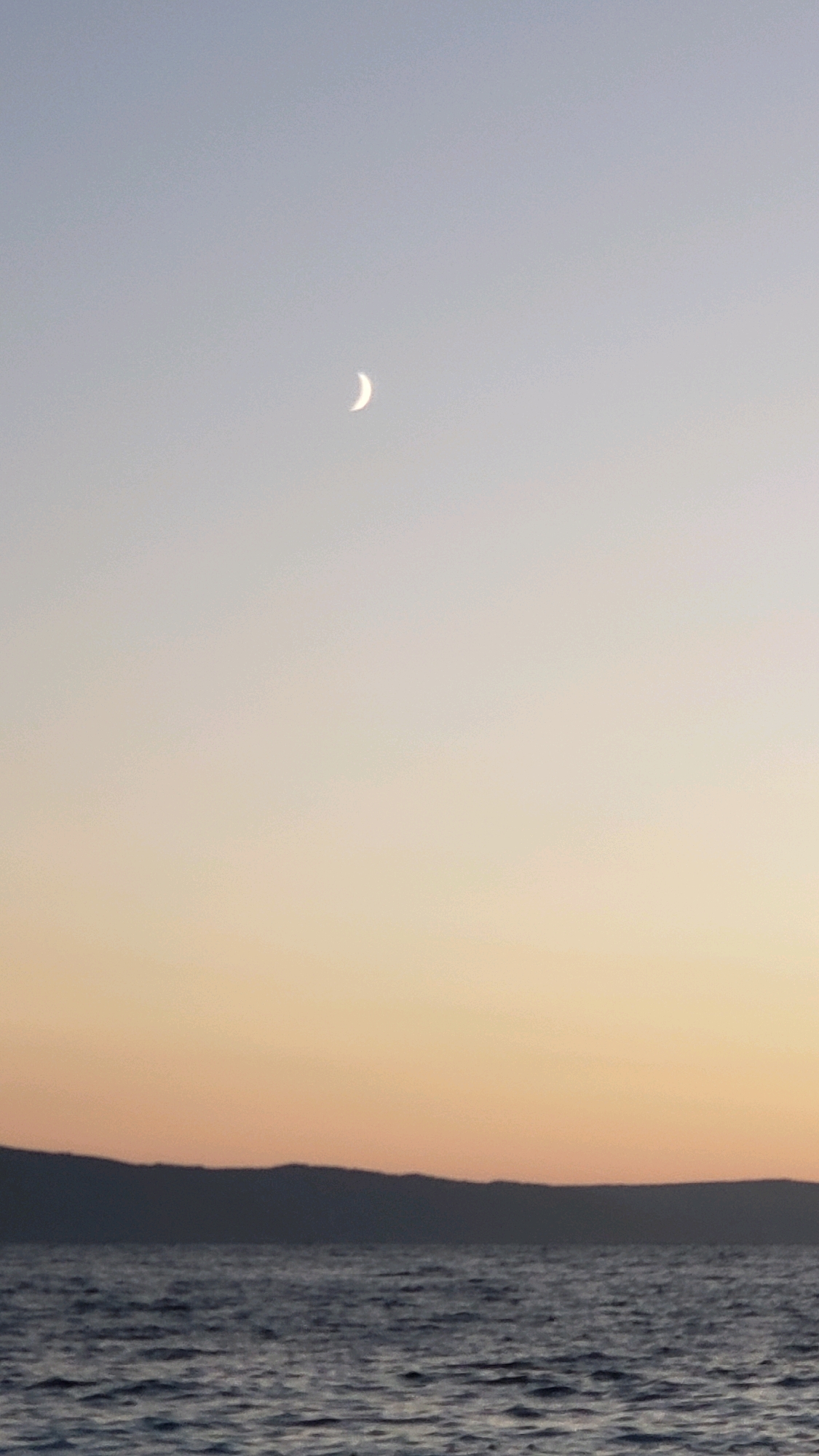A crescent moon is visible in the pale sky above a dark, silhouetted mountain range. The sky transitions from a light blue at the top to a warm orange hue near the horizon, indicating either sunrise or sunset. Below the mountains, a body of water with gentle ripples reflects the sky's colors. The scene suggests a tranquil coastal environment in Općina Tučepi, Croatia. There are no people or specific objects visible in the frame, besides the natural elements of the sky, moon, mountains, and water.