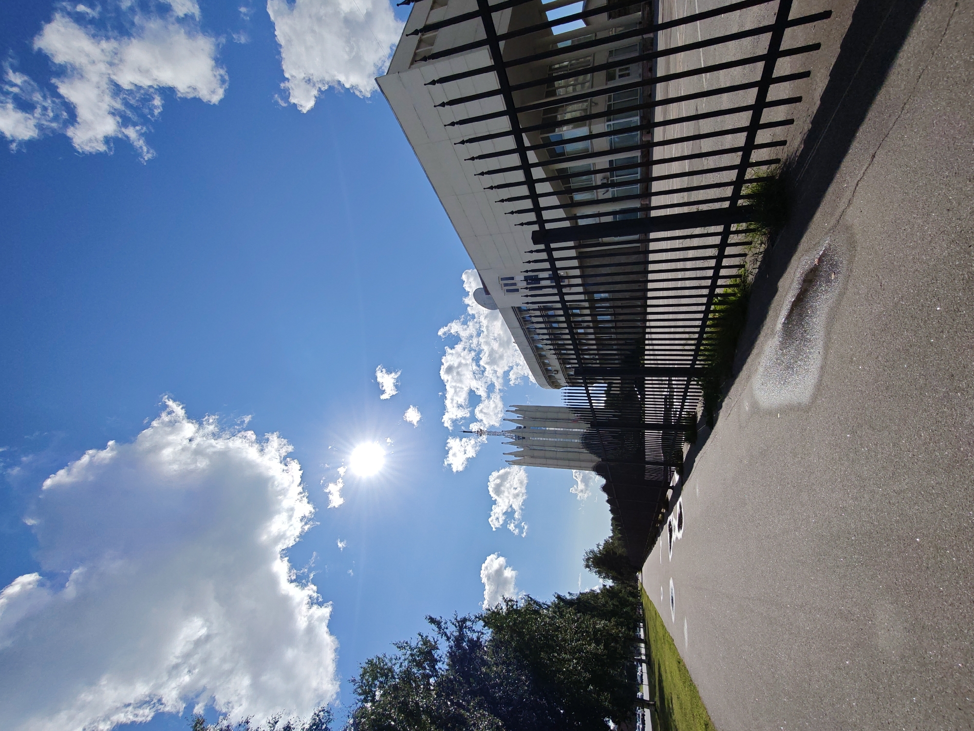 The image depicts a sunny day in Saint Petersburg, Russia, likely on the territory of Peter the Great St. Petersburg Polytechnic University. A wide, grey asphalt pathway runs diagonally from the bottom left to the middle distance, bordered on the right by a tall, black metal fence. Beyond the fence, a modern, light-colored building with large windows is visible, and further in the distance, a distinctive, tall, slender white building with a pointed spire topped with an antenna stands out against the bright blue sky. The sky is filled with scattered white clouds, and the sun is shining brightly, casting sharp shadows from the fence onto the pathway. To the left of the pathway, a line of lush green trees forms a border with a grassy area beyond. A few scattered puddles are visible on the pathway, reflecting the sunlight. No people are present in the scene.