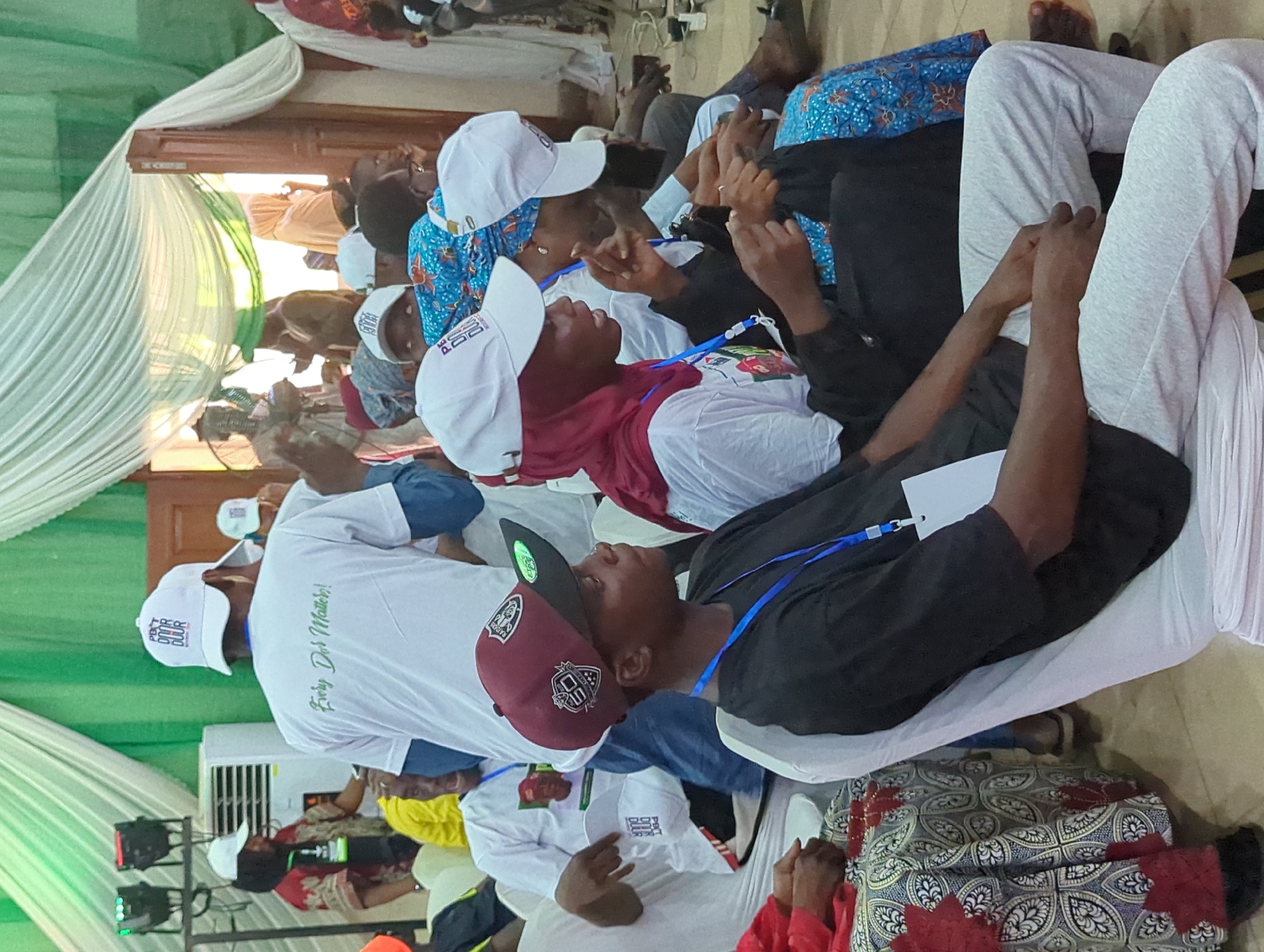 A medium-angle shot depicts an indoor gathering in Ungwan Liman, Nigeria. A group of seated individuals, likely attendees of an event, occupy chairs draped in white fabric. Many wear white baseball caps. One individual sports a maroon baseball cap and a black shirt. A white air conditioning unit is behind a man wearing a white shirt with the slogan "Every Dot Matters!". A green and white tent-like structure is in the background, adding to the event-like atmosphere.