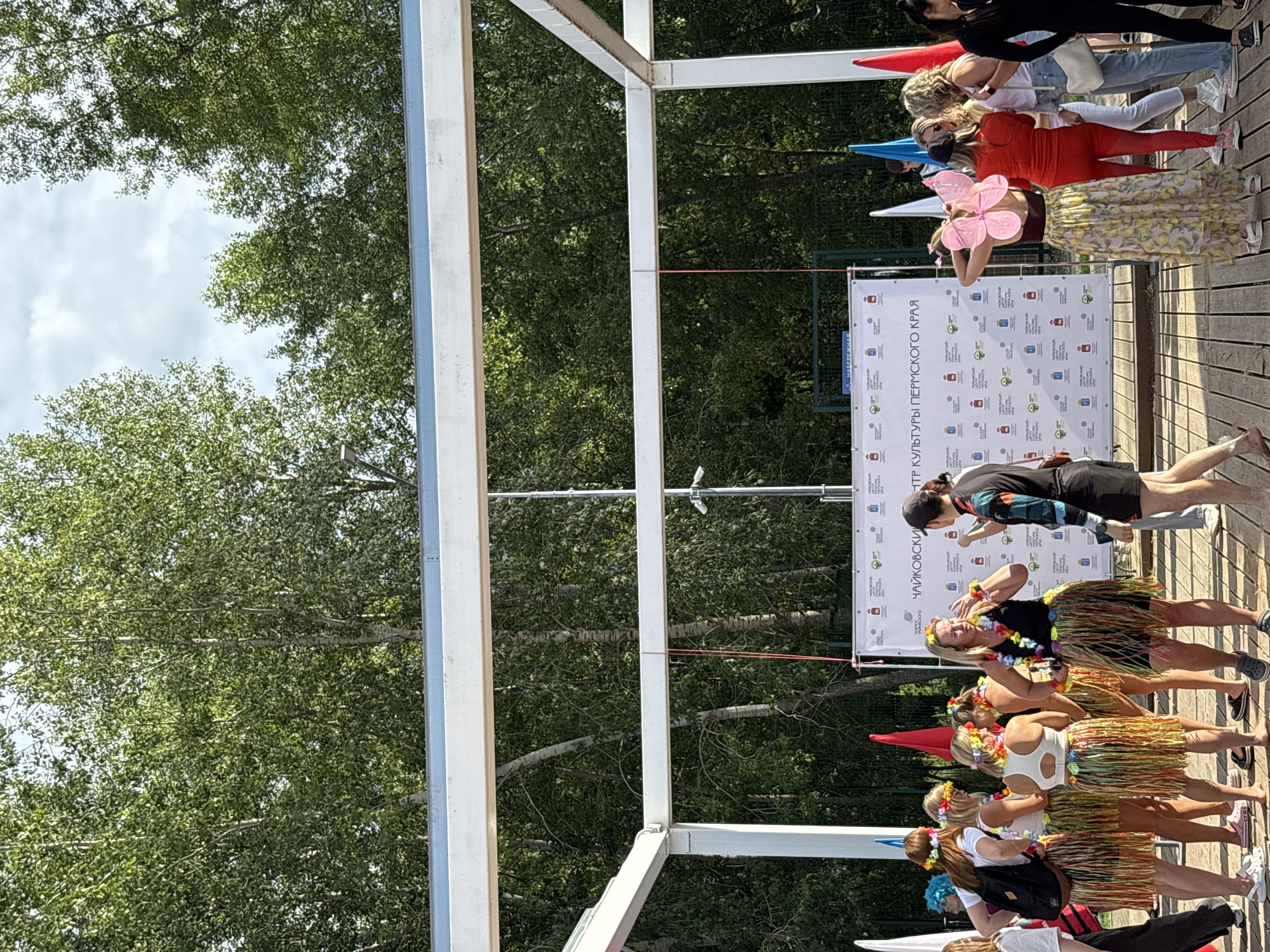 A group of people, mostly women dressed in Hawaiian-themed attire, stand gathered in front of a white banner with printed logos and text. The banner is displayed on a wooden platform, which appears to be outdoors. Trees with green foliage form a dense background, suggesting a park or natural setting. Some individuals are wearing leis and grass skirts, indicating a festive atmosphere, possibly a luau or cultural event. In the distance, more people are visible, along with colorful flags and a white canopy structure. The banner text includes Russian characters and mentions "Чайковский" and "Пермского края," indicating the location is Chaikovsky, Perm Krai, Russia. The overall scene suggests a community event or celebration taking place in an outdoor venue.