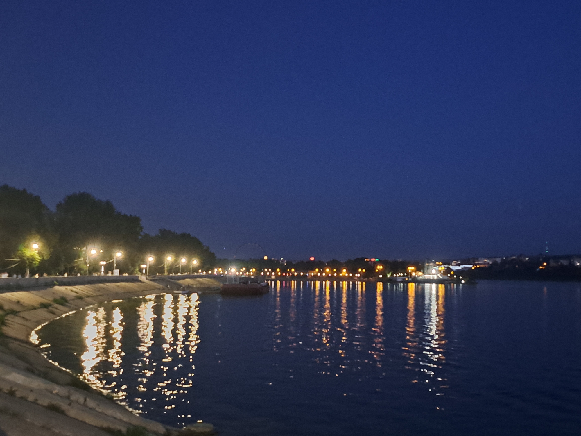 The image depicts a nighttime scene of the Angara River embankment in Irkutsk, Russia. The sky is a deep, clear blue. Along the riverbank, a paved walkway is illuminated by numerous streetlights, casting reflections on the dark water. Trees line the walkway, their silhouettes visible against the lights. On the far side of the river, a cityscape is illuminated with various lights, including a Ferris wheel and buildings. A small boat is visible in the water near the embankment. The reflections of the lights on the river create a shimmering effect.