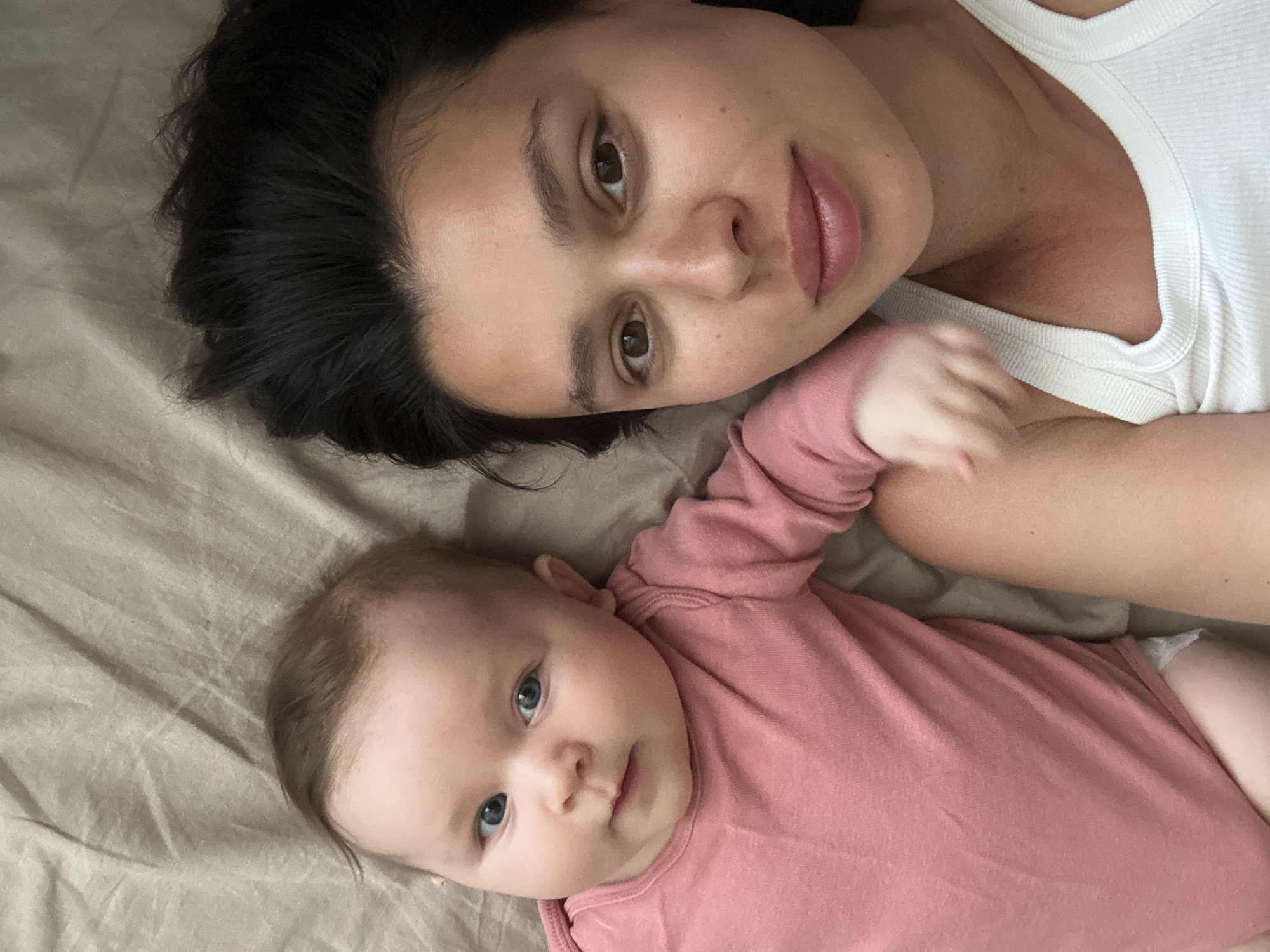 A woman and a baby are lying on a beige sheet. The woman has dark hair, brown eyes, and is wearing a white tank top. The baby has light brown hair, blue eyes, and is wearing a pink shirt. The baby is lying with its left arm outstretched. The setting is indoors, likely a bed. The image provides no information regarding the city of Chaikovsky, Russia.