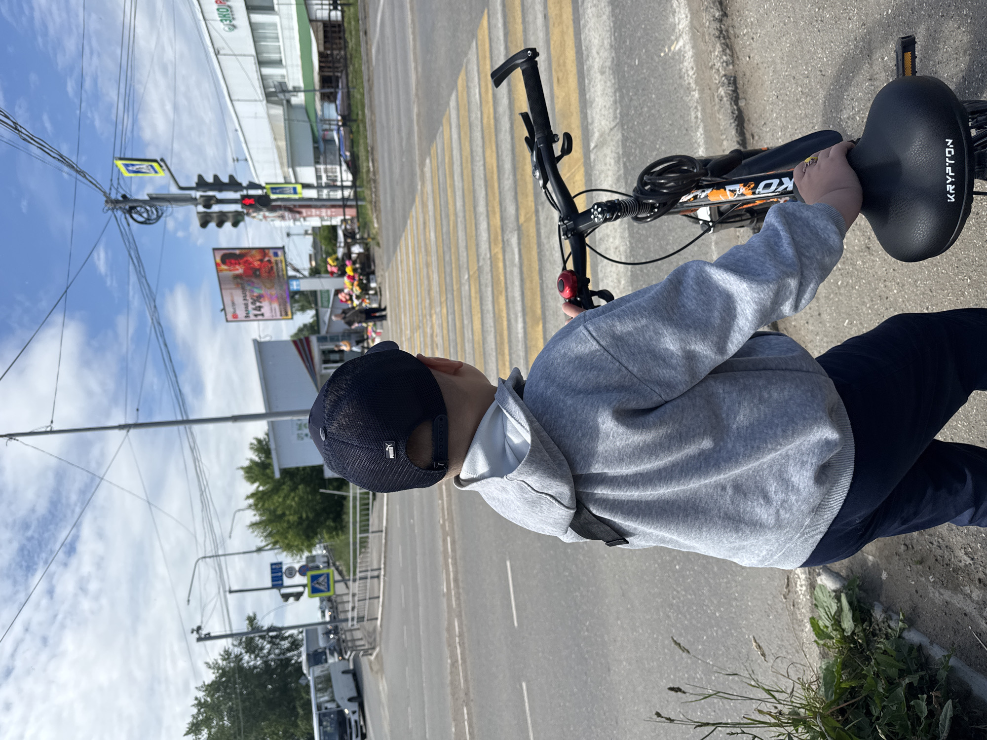 A young boy, observed from behind, stands on a paved sidewalk beside a yellow and white striped pedestrian crossing at an intersection in the city of Chaikovsky, Russia. He wears a navy blue Puma baseball cap, a grey hooded sweatshirt with a light grey inner hood, and dark blue trousers, holding onto a black bicycle with orange accents. His left hand rests on the black "KRYPTON" branded bicycle seat.
The child is positioned before the pedestrian crossing, facing a multi-lane asphalt road. A traffic light pole in the mid-ground displays a red light for vehicular traffic, accompanied by a blue square sign depicting a white walking person and a yellow diamond-shaped sign with a black walking person, both indicating a pedestrian crossing. A white stop line is visible on the road before the crossing.
In the background to the left, several white buses are in motion on the road. Across the intersection, various white commercial buildings line the street. One building facade displays partial Cyrillic text, including "ЭКО Р", while another features a large billboard showing an advertisement with Cyrillic text and the number "14%". A street vendor's stall with an array of colorful flowers is situated on the opposite sidewalk. The scene is illuminated by bright sunlight under a partly cloudy sky, indicating daytime. Multiple utility poles and overhead wires crisscross the sky. Weeds and grass grow along the edge of the sidewalk in the foreground.