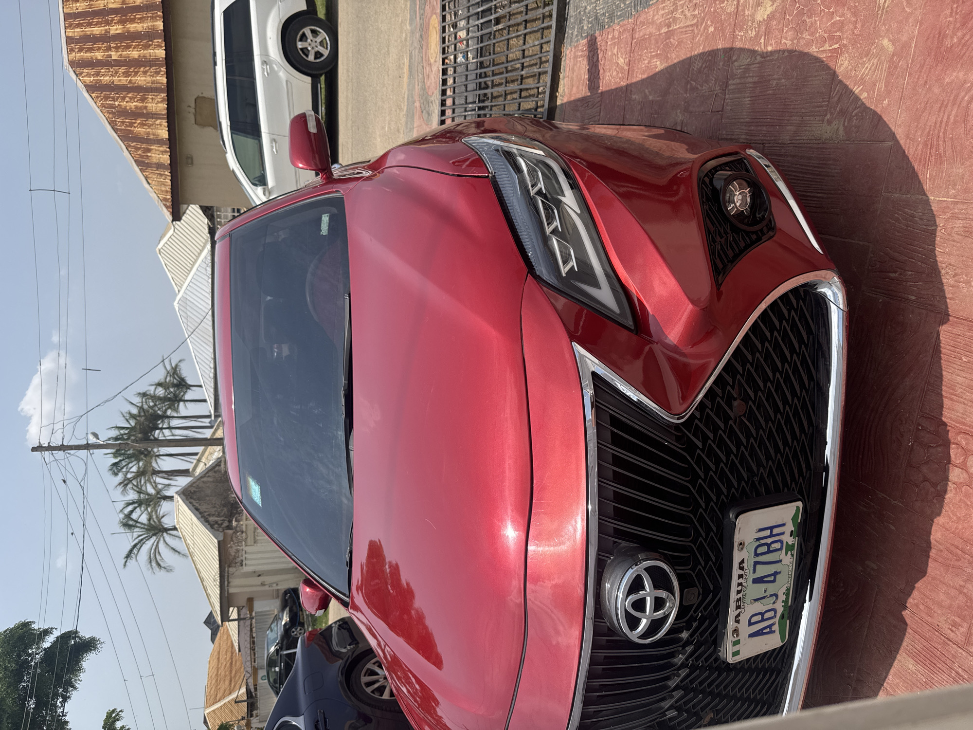 This image shows the front of a red Toyota sedan parked outdoors in what appears to be a residential or commercial area in Abuja, Nigeria. The car is the primary subject, with its grille, headlights, and hood clearly visible. A license plate is attached to the front grille, displaying the text "ABJ 47BH" and "ABUJA CENTRE OF UNITY".
In the background, a white SUV is partially visible, suggesting other vehicles are present. The setting includes a building with a corrugated metal roof and some palm trees, indicating a tropical or subtropical climate. The ground is paved with reddish-brown textured material. Sunlight casts strong shadows, suggesting it is daytime, likely midday. The sky is clear and blue. The overall scene depicts a common urban or suburban environment in Abuja. There are no people visible in the image.