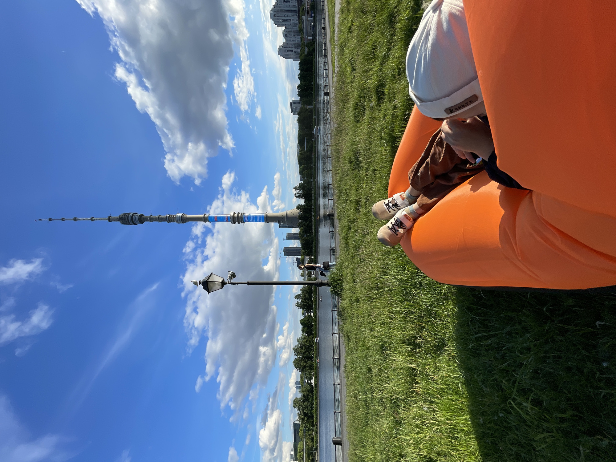 A person wearing a white hat and brown clothing is sitting in an orange inflatable lounger on green grass. In the background, there is a cityscape including the Ostankino Tower, a body of water, and a street lamp. The location is in the city of Unknown, Unknown.