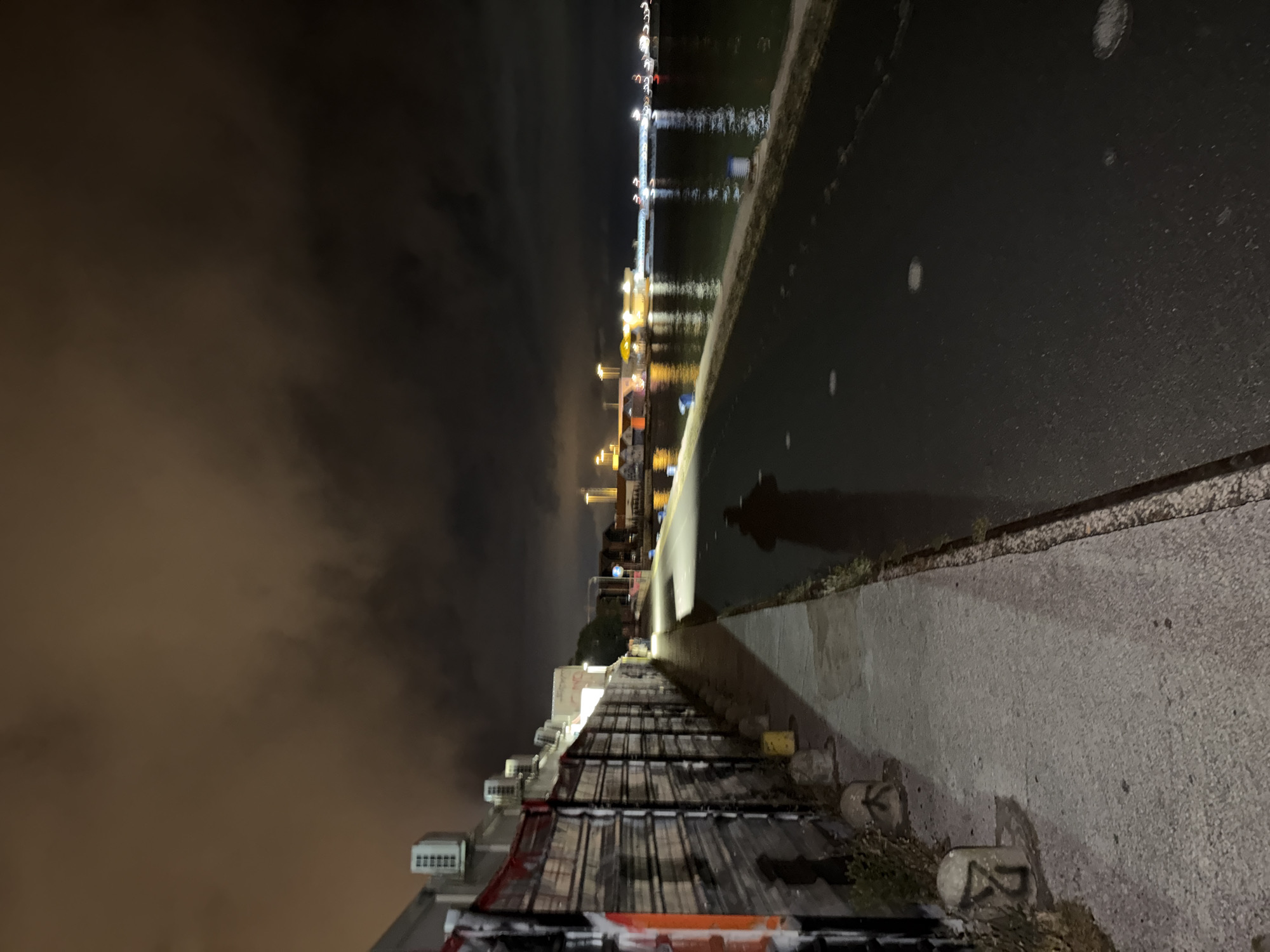 Captured at night in Lisbon, Portugal, the image presents a perspective along a waterfront walkway. The walkway, constructed of concrete, is adjacent to a narrow road. A line of barriers, painted with graffiti, separates the walkway from a building to the left. To the right of the road is a body of water, reflecting the lights of buildings and structures across it. The overall setting is urban, with street lighting illuminating the scene. The sky is obscured by a heavy fog. A human shadow is seen cast on the walkway from the street.