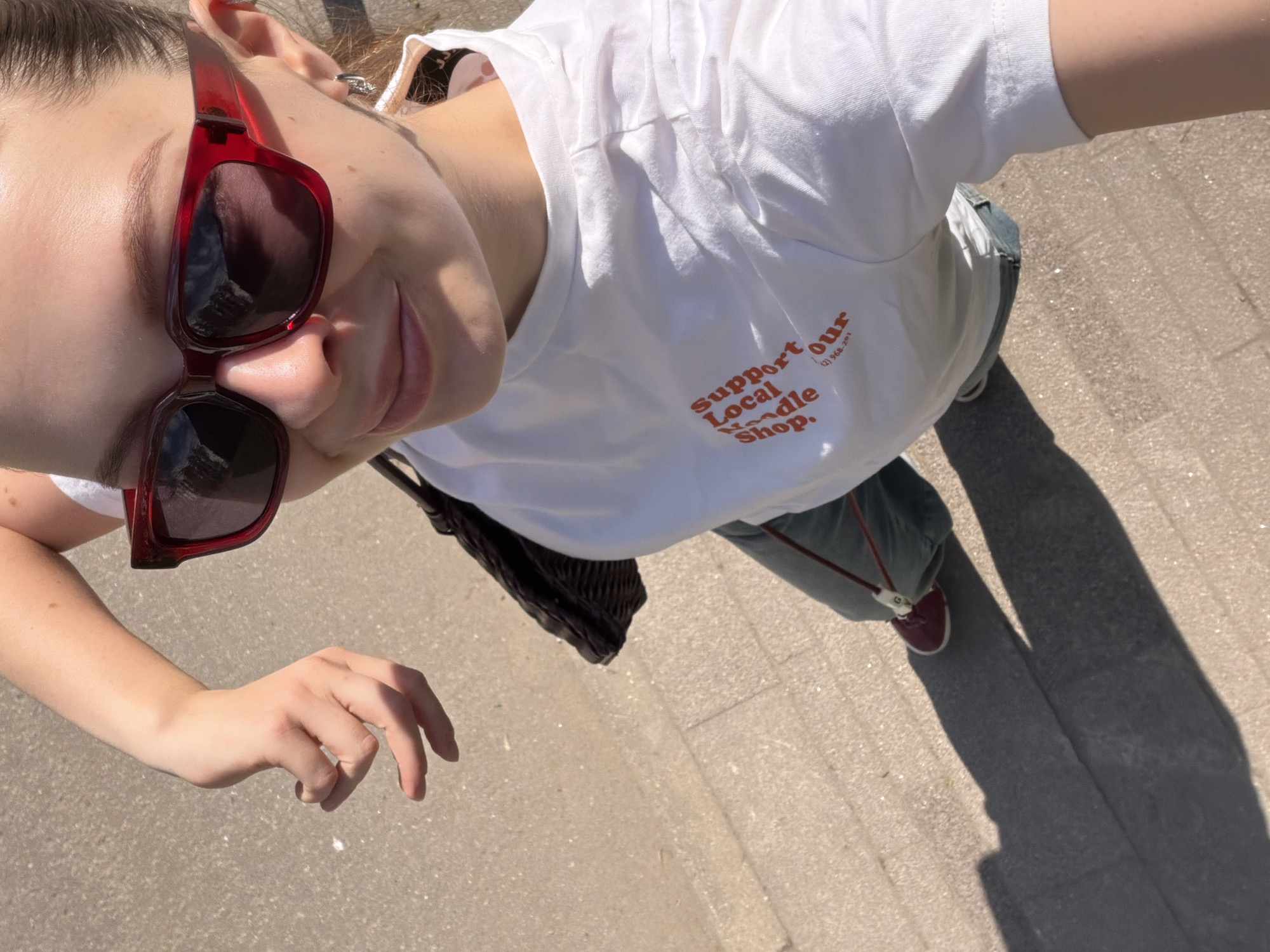 A person, likely female, is standing on a concrete sidewalk, angled downwards in a self-taken photograph. The person is wearing red sunglasses and a white t-shirt printed with "support our Local Noodle Shop." They are also wearing green pants and red sneakers. A purse is visible on their right side. The scene takes place outdoors in bright sunlight. No information is available to identify the city of Khimki, Russia.