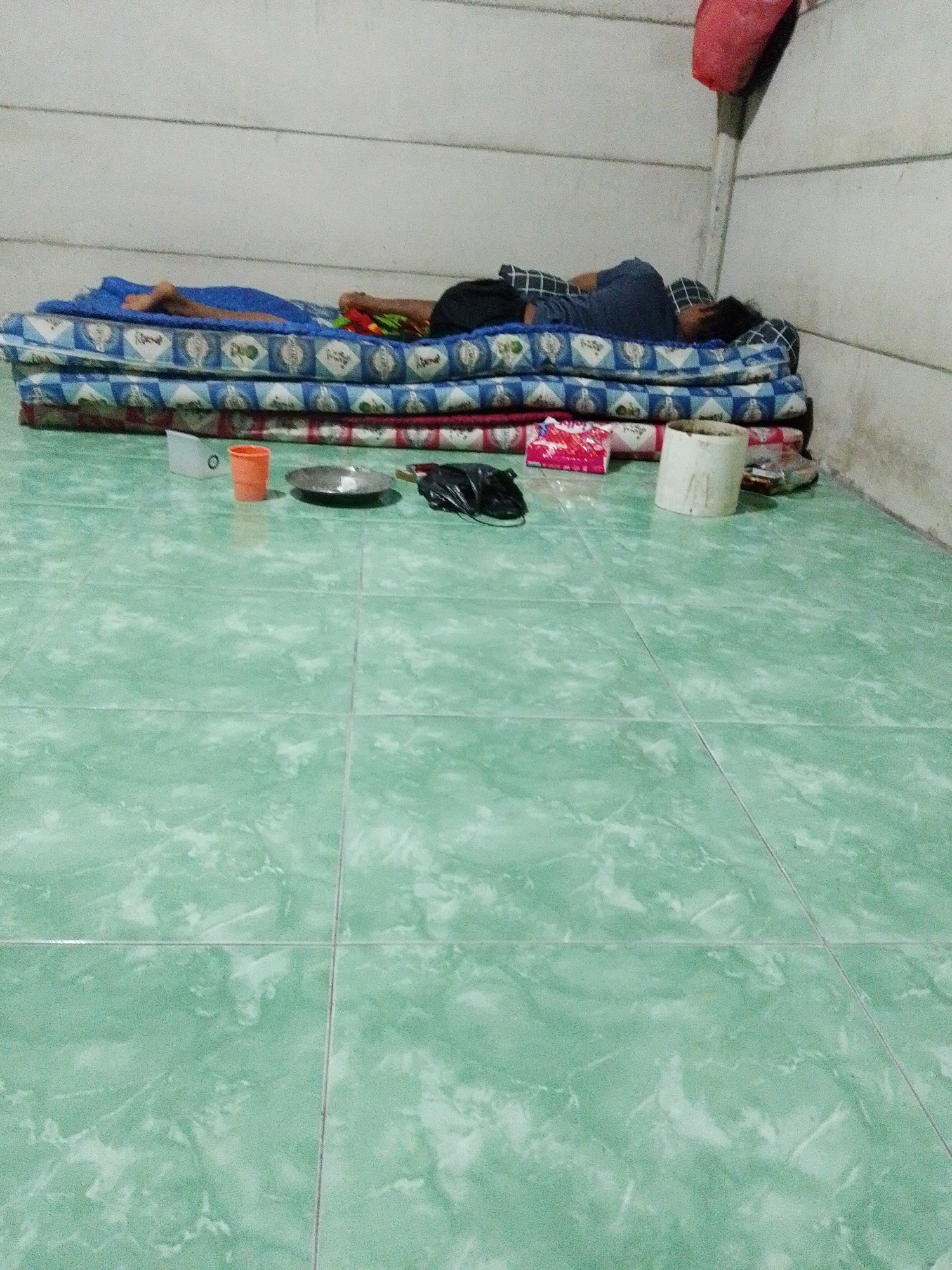 A person is asleep on a stack of mattresses in a room in Bukit Ketok, Indonesia. The room has pale, textured walls and green tiled floors. An orange cup, a metal dish, a pair of black shoes, a bag of candy, a roll of duct tape, and miscellaneous trash are arranged on the floor near the mattresses. The person is wearing a dark blue shirt and black shorts.