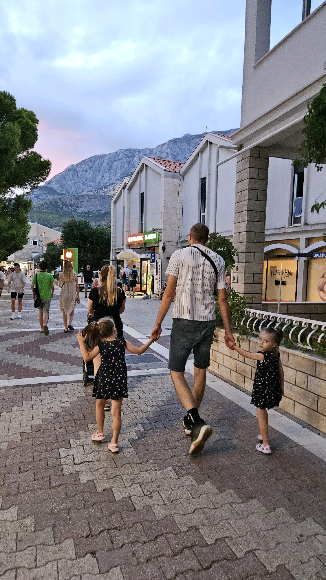 A man and two young girls walk hand-in-hand along a paved pedestrian street in Općina Tučepi, Croatia. The man, wearing a striped t-shirt and denim shorts, holds the hands of two girls dressed in matching black floral-print dresses and pink sandals. The girls are walking in front of him, with the girl on the left appearing to push a stroller. Other pedestrians and buildings line the street. In the background, mountains rise under a twilight sky with hues of pink and blue. The street is paved with interlocking bricks, and a white railing with a decorative pattern runs along the right side of the walkway.