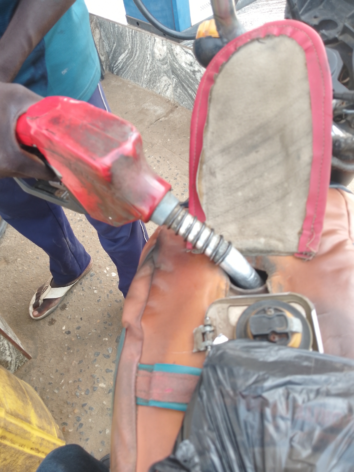 In Serti, Nigeria, a close-up shot captures a person in blue trousers and a teal t-shirt fueling a motorcycle. The red fuel nozzle is inserted into the motorcycle's fuel tank. The background shows a textured wall and a concrete ground. A black plastic bag is visible near the fuel tank. The scene appears to be at a fuel station.