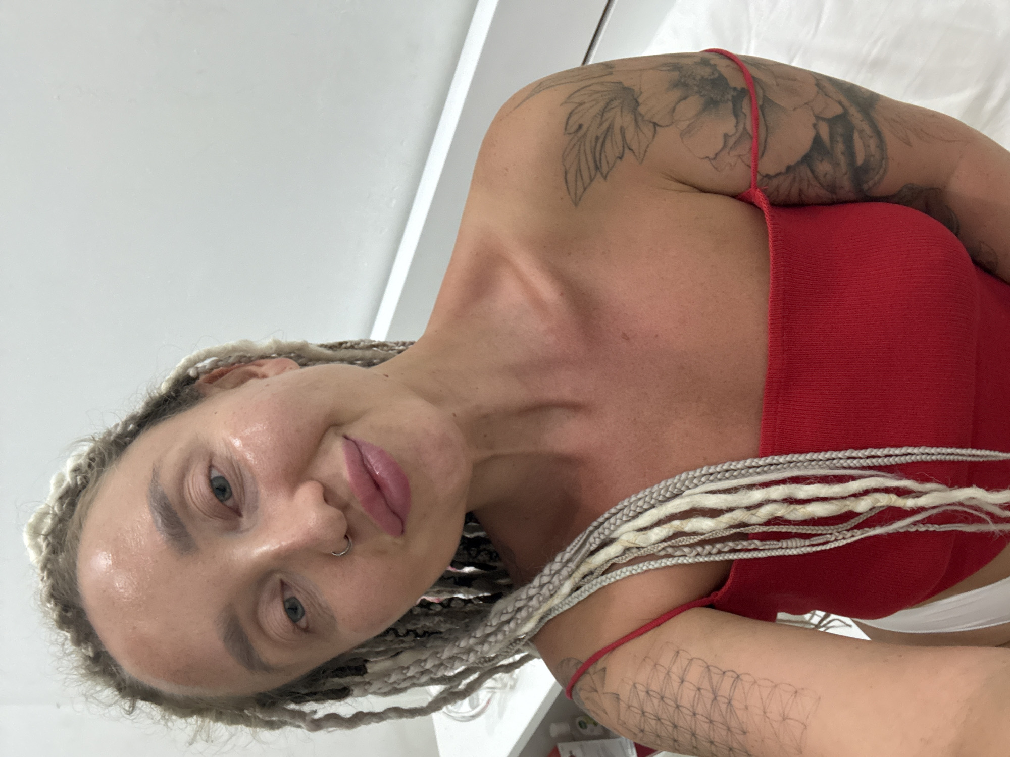 A woman with blonde dreadlocks and a nose ring poses for a selfie in what appears to be a dimly lit room, possibly a spa or bedroom. She is wearing a red tank top and has tattoos on her arms and shoulder. The background is mostly white, with some shelves and bottles visible on the left side of the image. The lighting is soft and highlights the woman's features.