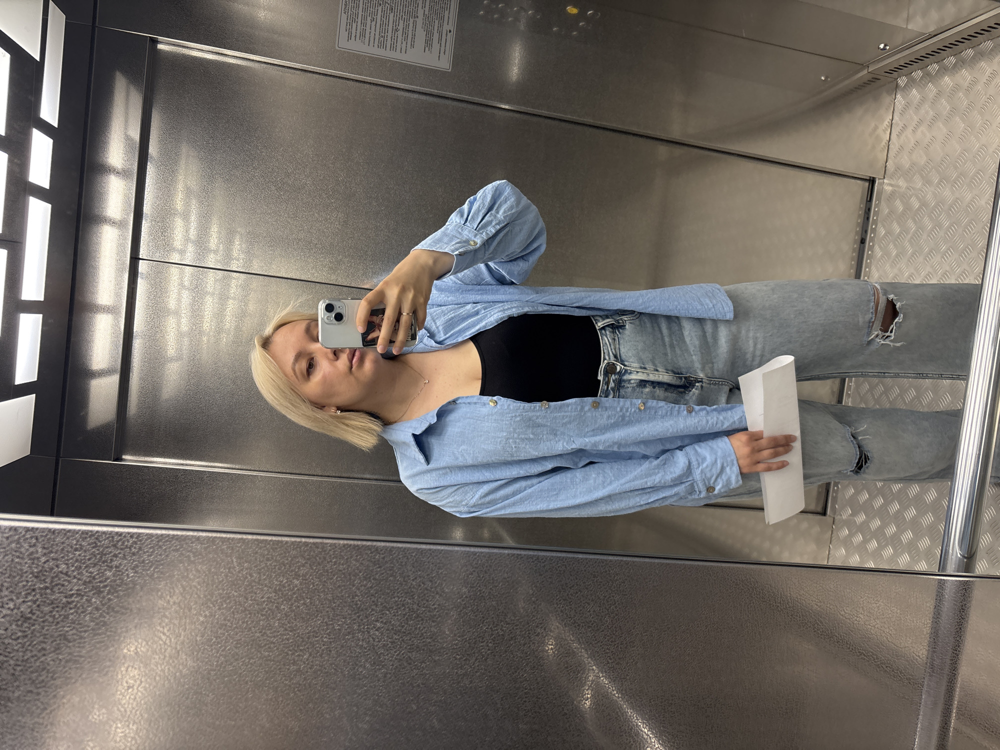In a modern elevator, a young woman with blonde, shoulder-length hair stands in front of a brushed metal wall. She is wearing a light blue, oversized button-down shirt over a black tank top and light-wash jeans with ripped knees. She holds a white folded paper in her left hand and is taking a selfie with her phone, which is partially visible in her right hand. The elevator's interior is characterized by stainless steel panels and a textured metal floor. The lighting appears to be from overhead fluorescent fixtures. The scene takes place in an elevator, potentially located in a building in Chaikovsky, Russia, given the context provided.