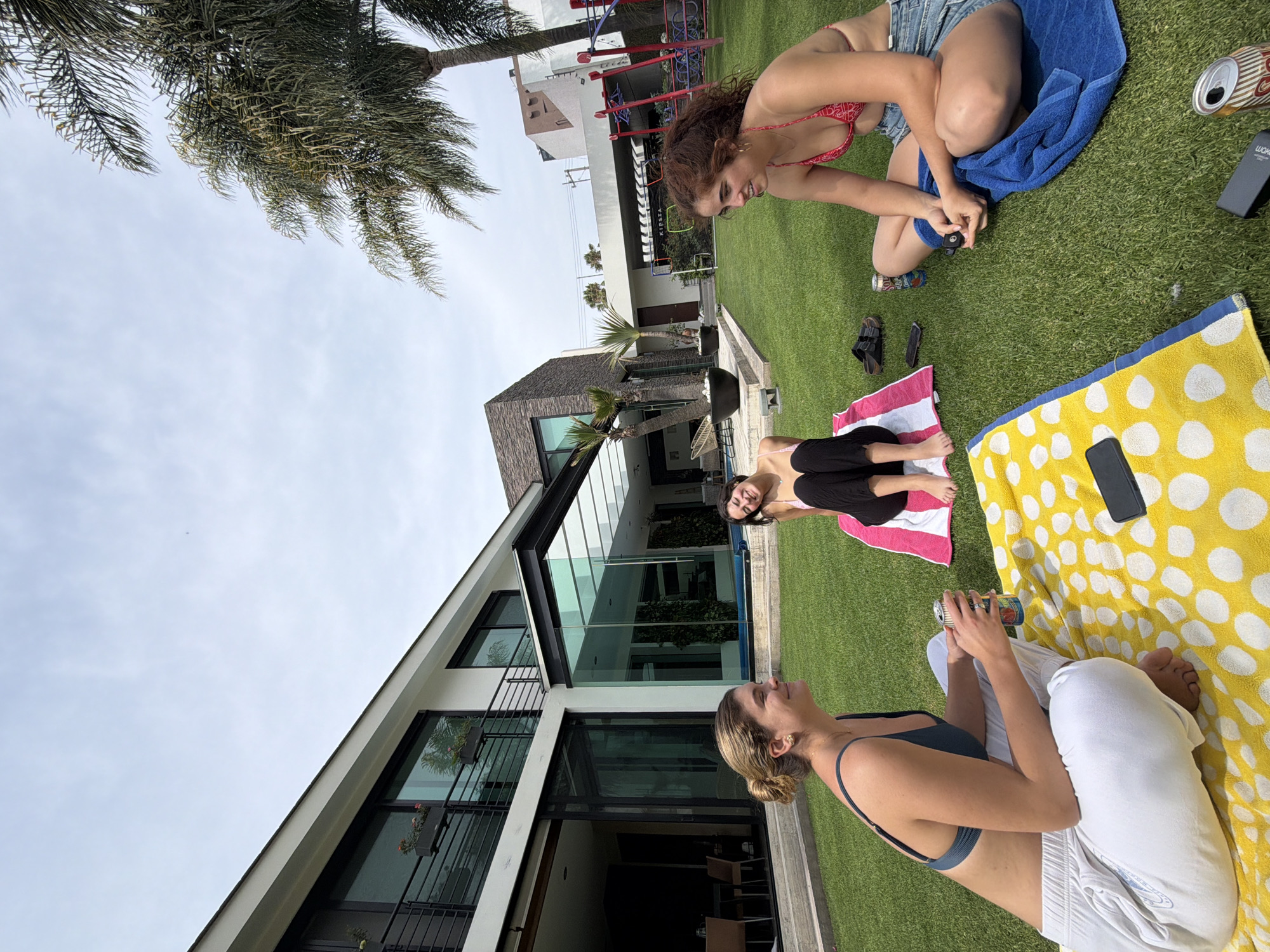 The image shows three women on a grassy lawn. One woman, wearing a red top and denim shorts, is sitting on a blue towel, with a can and a black phone next to her. Another woman is lying on a pink and white striped towel. The third woman, wearing a blue top and white pants, is sitting on a yellow and white polka-dot towel, holding a can. A modern house with large windows and a flat roof is in the background. A palm tree is partially visible in the upper left corner. In the upper background there is a building with playground equipment.
