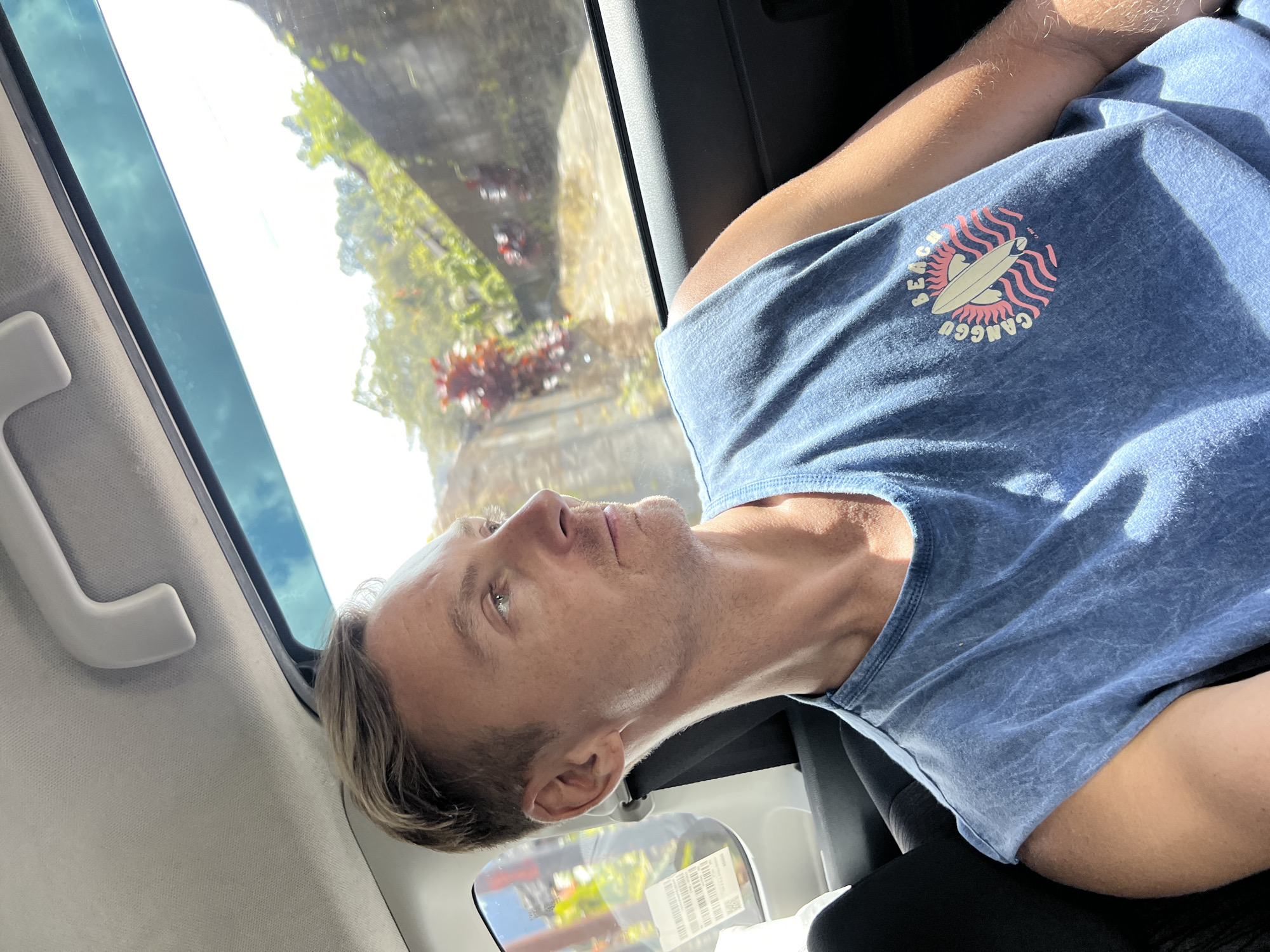 The image shows a light-skinned man with short blond hair sitting in a car. He is wearing a blue t-shirt with a graphic featuring a surfboard and the words "BEACH CANGGU." The car's window provides a view of the outside, including what appears to be a bridge or viaduct with greenery on the sides. The location is Baturiti, Indonesia. The man is gazing out the window. The interior of the car is visible, including the window frame, a grab handle, and part of the seat.