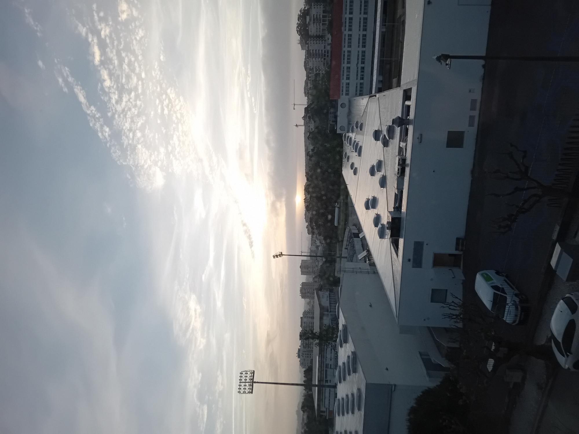 This landscape image depicts an urban or suburban environment under a bright, low sun, suggesting either sunrise or sunset. The sky is filled with soft, diffused clouds, with the sun appearing as a brilliant orb near the horizon, creating a warm, hazy glow.
In the foreground and mid-ground, numerous buildings are visible. On the right, a large, modern building with a flat roof features several circular vents or skylights. Below it, two white vehicles, one with green detailing, are parked on a dark paved area. Bare trees and other vegetation are scattered throughout.
Further into the scene, a mix of residential and commercial buildings stretches across the horizon, some with distinct red-tiled roofs, appearing silhouetted against the bright sky. A tall stadium-style light tower with multiple lamps is prominent on the left side of the frame. Another street light pole is visible closer to the foreground. In the distance, a construction crane can be seen, indicating ongoing development.
The time of day is clearly dawn or dusk due to the low sun and soft lighting. The weather appears partly cloudy with clear visibility. On one of the buildings in the middle distance, the word "SUPERMARCHÉ" is faintly visible.