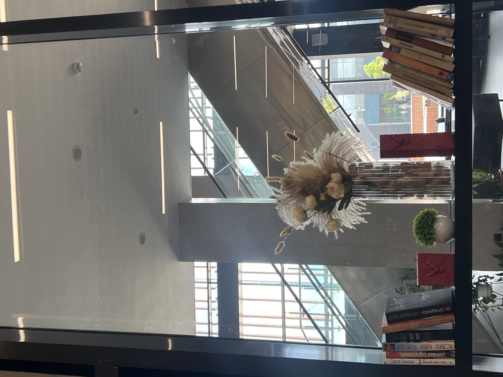 The image shows an interior view of a modern building, likely a library or office space, with a focus on a shelf containing books, decorative items, and a vase of dried flowers. The shelf is positioned in front of a large window, which reveals an escalator system and the exterior of a building with architectural elements characteristic of contemporary urban design. The scene is indoors, with artificial lighting present, including linear fluorescent lights on the ceiling. The shelf itself is black and appears to be made of metal. On the left side of the shelf, there are several books standing upright, their spines displaying text in Cyrillic. To the right of these books, there are two red decorative candles, one with an embossed image of a deer, and a small potted green plant. Further to the right, a tall, clear glass vase contains a dried floral arrangement, predominantly featuring pampas grass and some light-colored artificial flowers. The background shows the interior architecture of the building, with polished concrete pillars and the structure of the escalator. The exterior view through the window suggests a daytime setting, with daylight illuminating the scene. The specific location in Veliky Novgorod, Russia, is not explicitly provided in the visual information, but the presence of Cyrillic text on the book spines indicates a Russian-speaking environment.