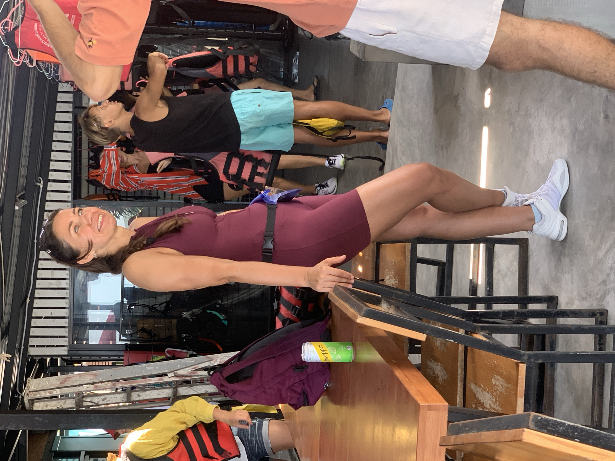 In Ko Samui District, Thailand, a woman stands to the right of a wooden table, wearing a maroon athletic outfit and white sneakers. Her hair is pulled back in a braid. To her left, a purple backpack rests on the table next to a can of Schweppes. In the background, several life vests and other gear are visible, suggesting an outdoor activity center or rental shop. Other individuals are present in the background, some wearing life vests. The environment appears to be an open-air structure with concrete flooring and hanging overhead equipment.