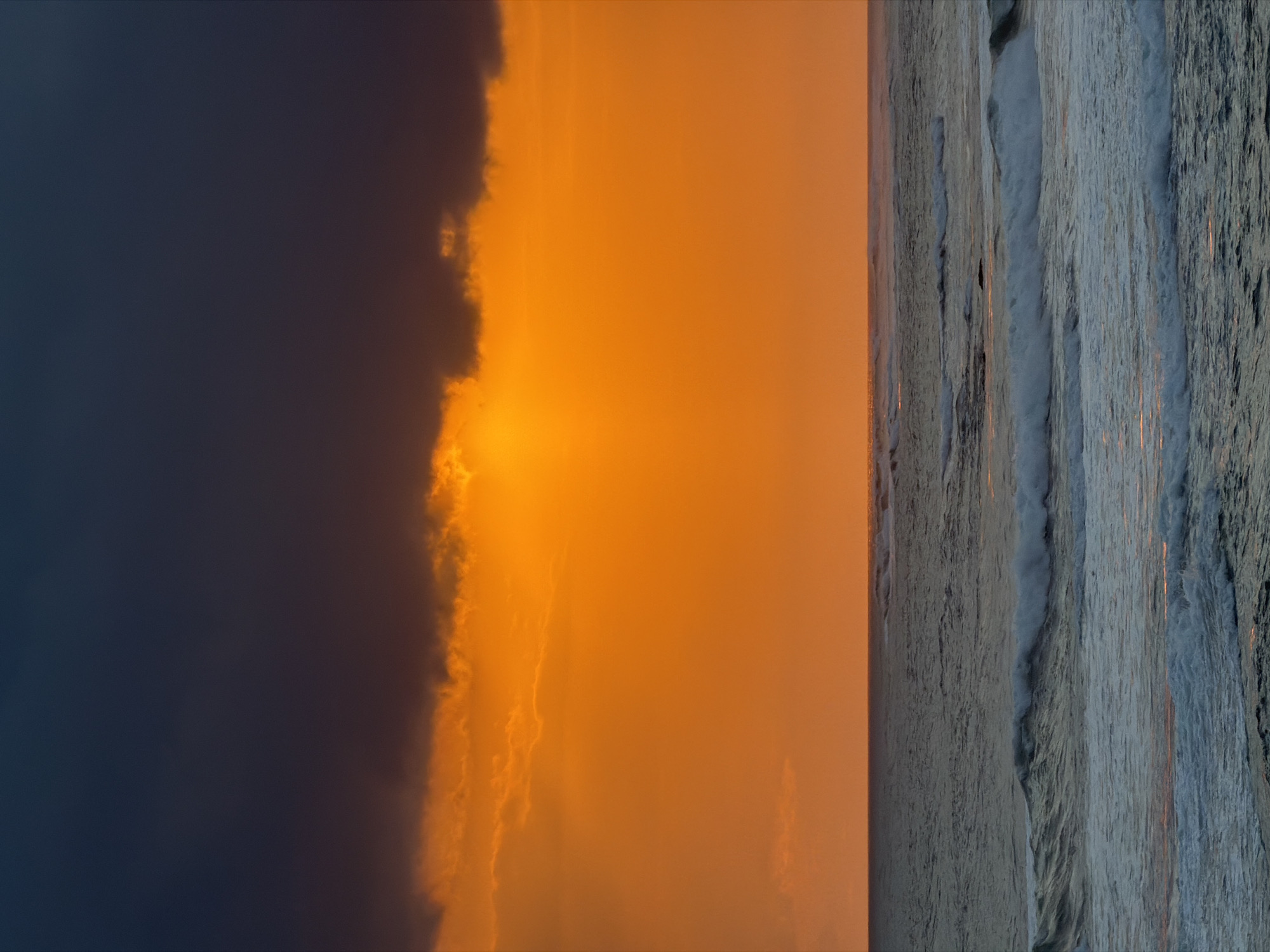 The image depicts a dramatic sunset over the ocean. The sky is divided into two distinct sections: the upper portion is a dark, brooding gray, while the lower section is ablaze with a vibrant, fiery orange. This intense orange hue illuminates a band of clouds near the horizon, creating a striking contrast with the darker sky above. Below the sky, the ocean stretches out, appearing gray and turbulent, with waves breaking and cresting with white foam. The overall impression is one of raw natural beauty and a powerful atmospheric display. The location is identified as Mengwi, Indonesia, suggesting a coastal scene. There are no people, subjects, or specific objects clearly identifiable in the image beyond the natural elements of sky and sea.