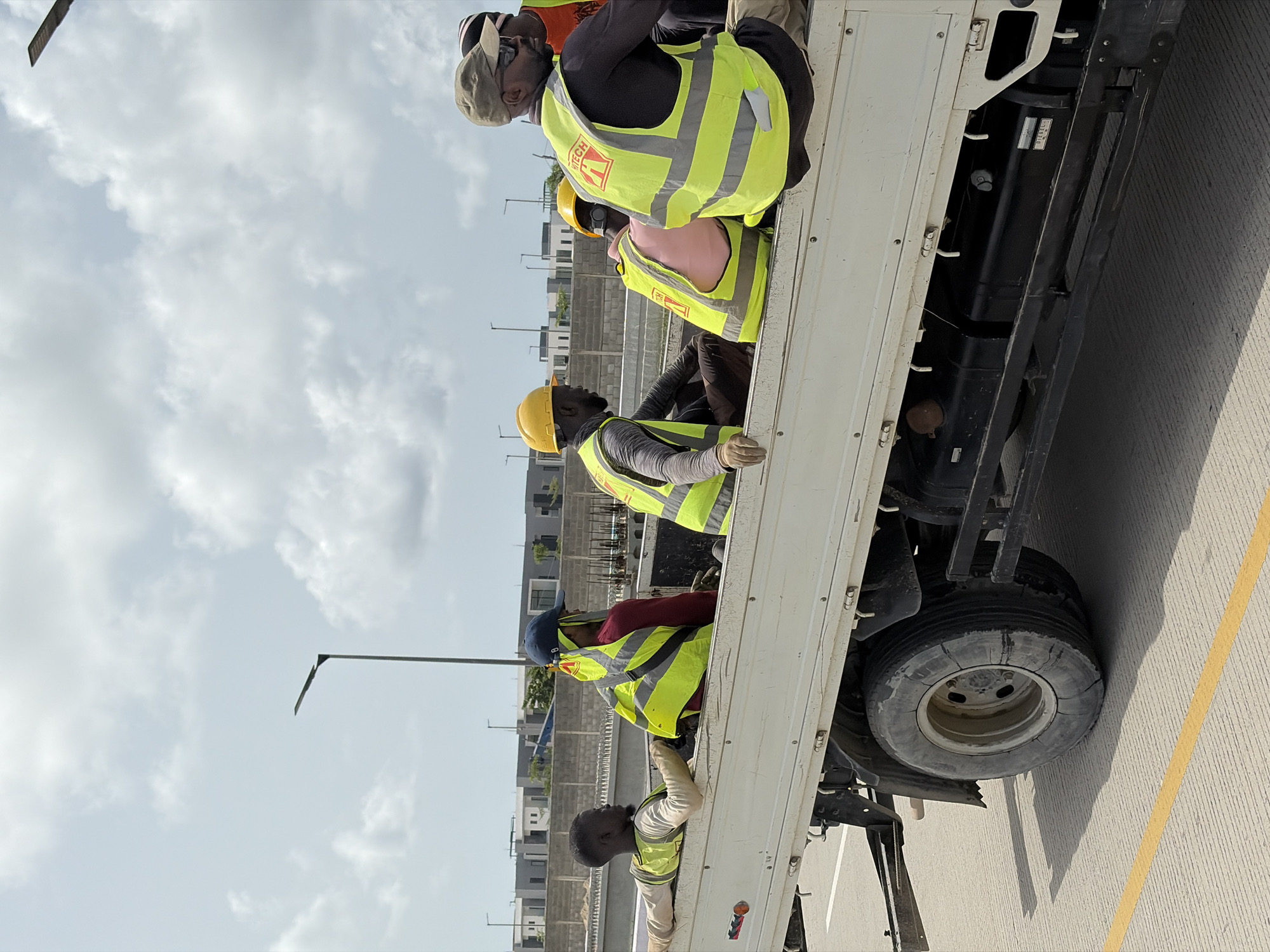 The image depicts several workers in Igboefon, Nigeria, riding in the back of a truck. They are wearing hard hats and high-visibility vests. The truck is traveling on a paved road. Buildings are visible in the background. The sky is cloudy.