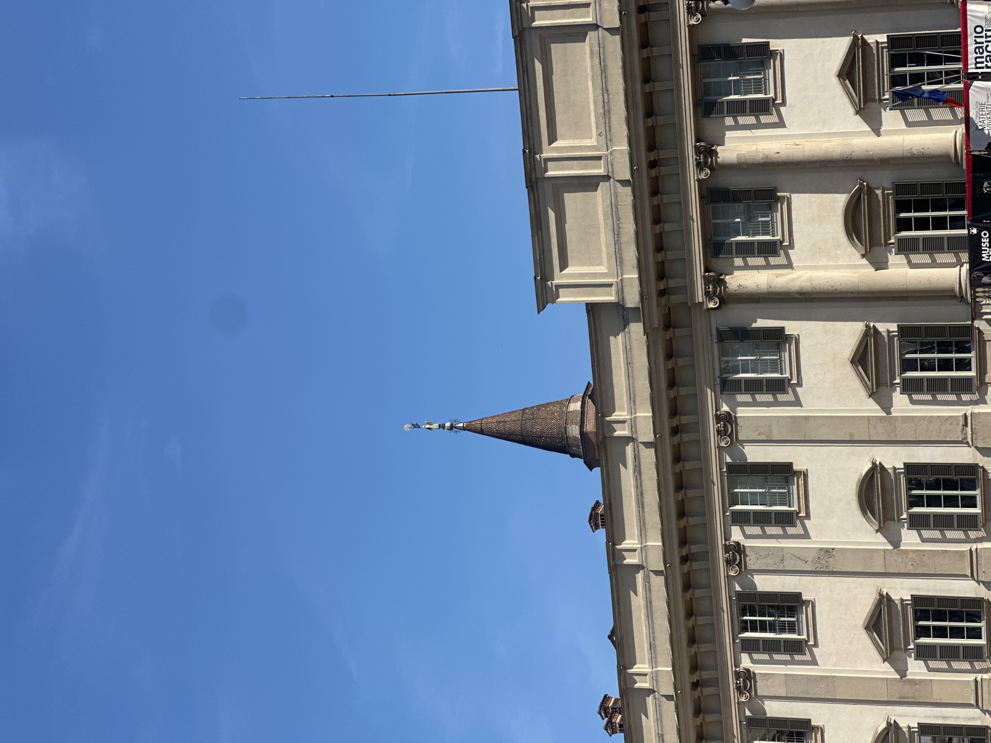 A bright blue sky with a few wispy clouds forms the background for a historic building in Milan, Italy. The building features a pale beige facade with classical architectural details, including ornate columns, cornices, and rectangular window frames. Many of the windows have dark shutters, some of which are partially open, revealing glimpses of their interiors.
A prominent conical spire with a tiled roof rises from the building, topped with a small, decorative finial. To the right of the spire, the building's facade continues upwards, showcasing multiple rows of windows. Several banners are visible on the right side of the building, one of which displays text that includes "mario raci" and "MUSICO". The sunlight casts strong shadows on the building's facade, indicating it is daytime, likely mid-morning or afternoon. The scene evokes a sense of historical grandeur and urban exploration.