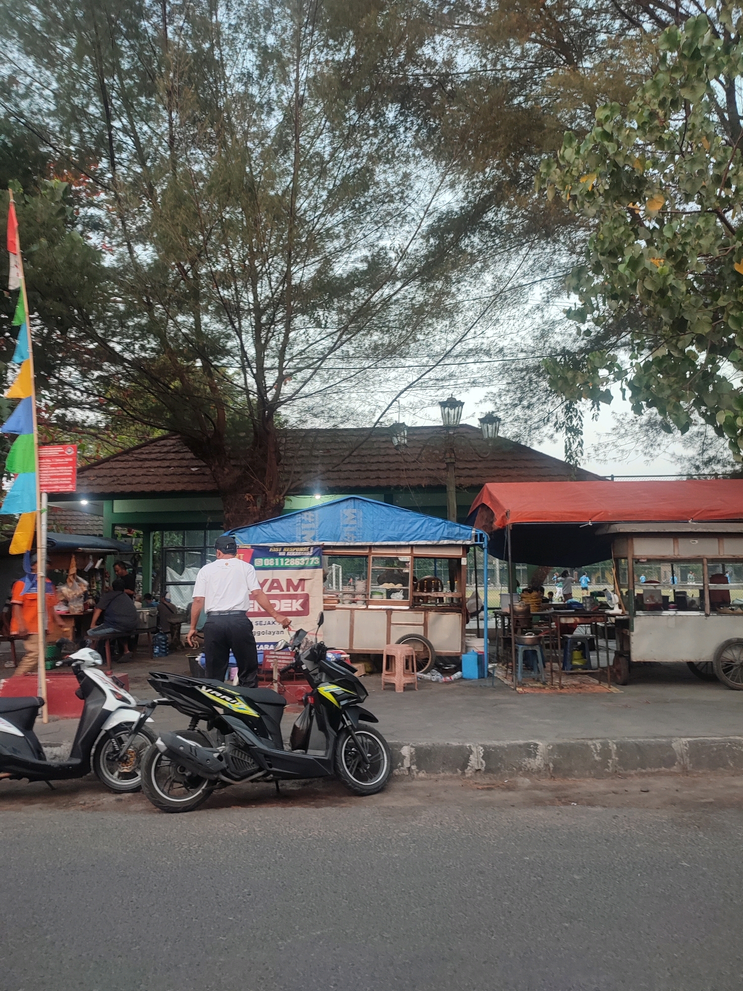 The image depicts an outdoor scene in Yogyakarta, Indonesia, likely near a public space such as a park or sports field, given the presence of people engaged in activity in the background. In the foreground, a paved road is visible, with a raised concrete curb separating it from a pedestrian walkway. Several motorcycles are parked along the walkway.
On the walkway, there are food stalls set up. One stall has a blue canopy, and another has a red canopy. Both appear to be mobile food carts. A man in a white t-shirt and dark pants is standing near the blue-canopied stall, facing away from the camera. He is wearing a black cap and appears to be interacting with the stall or its surroundings.
Behind the stalls, a building with a brown tiled roof is visible, possibly a pavilion or a small structure. Further in the background, several individuals are seen, some wearing blue shirts, potentially involved in a sports activity on a grassy area. The overall atmosphere suggests a casual, communal gathering place, likely during the late afternoon or early evening, with the trees suggesting a verdant environment. The presence of flags on a pole adds a touch of local festivity or identification.