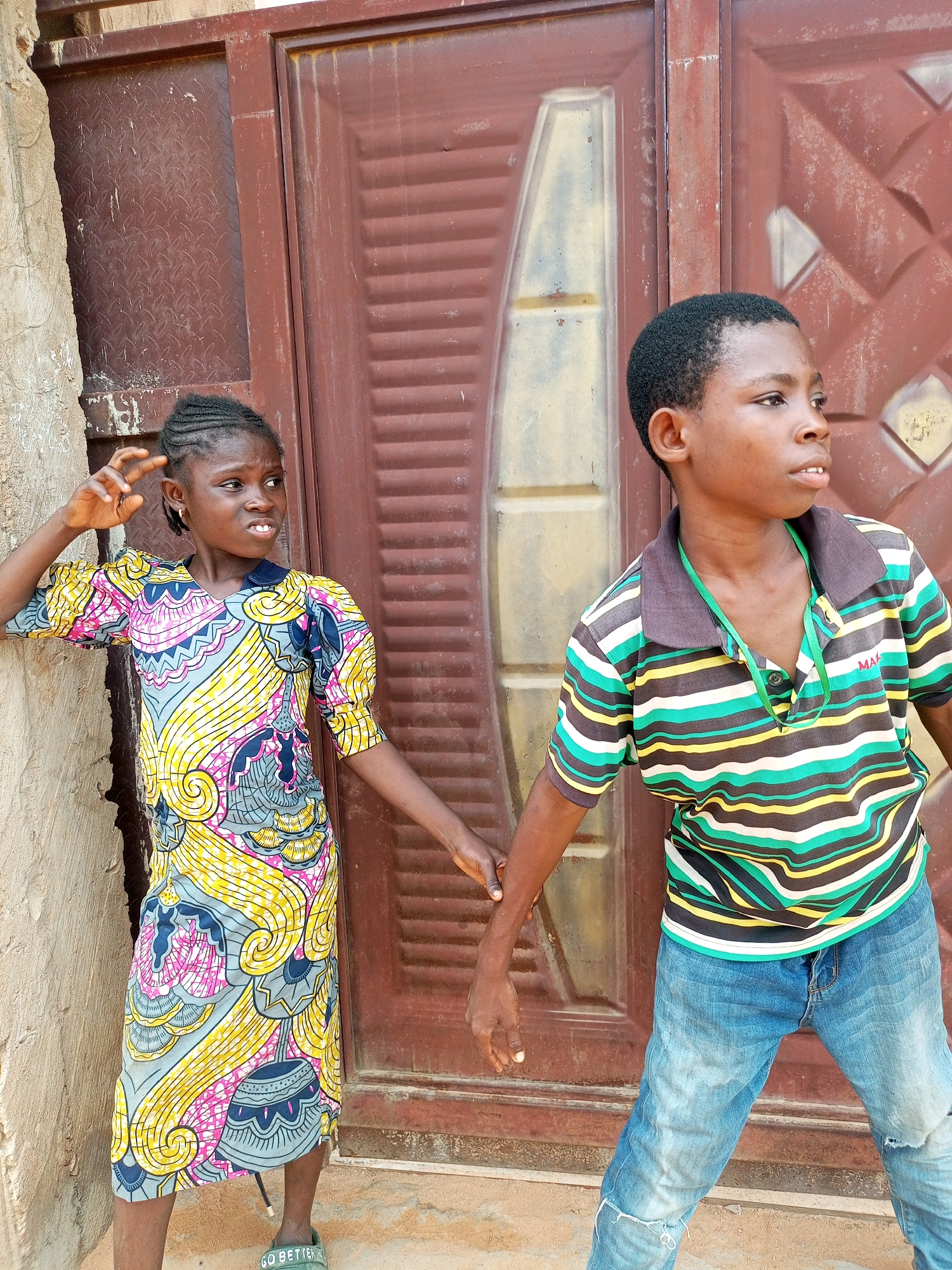 A medium shot captures a young Black girl and boy standing in front of a brown metal door in what appears to be an urban environment in Tempure, Nigeria. The girl, positioned on the left, wears a vibrantly patterned dress with puff sleeves in shades of yellow, pink, and blue. Her hair is styled in braids, and she is looking off to the side with her right hand raised near her head. The boy, on the right, is dressed in a striped polo shirt featuring green, yellow, white, and black bands, paired with distressed blue jeans. He is looking to his right, his gaze fixed on something out of frame. Their hands are clasped, suggesting a connection between them. The background consists of a textured, brown metal door with a decorative pattern and a beige, rough-textured wall to the left. The ground is a dusty, reddish-brown surface.