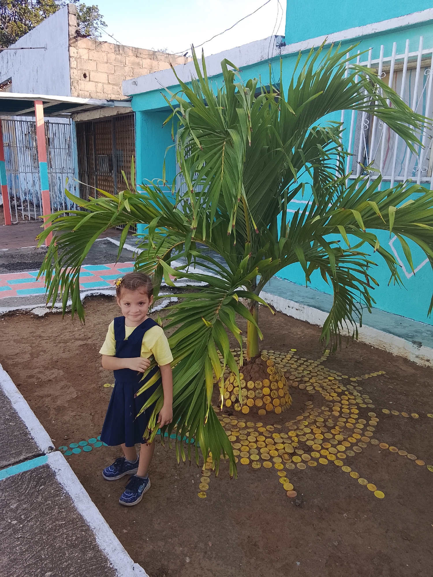 A young girl stands next to a small palm tree in Altagracia, Venezuela. The girl wears a yellow t-shirt and a dark blue dress with blue shoes. Her hair is adorned with a yellow flower clip. The palm tree's base is surrounded by dirt decorated with yellow circular objects, perhaps coins, arranged in a radiating pattern. Behind the tree is a turquoise building with white window bars and trim. Adjacent to the turquoise building is a dilapidated structure with a partially visible gate, painted in multiple colors. A sidewalk with painted square shapes runs along the lower left side of the frame. The ground is dirt, and the sky is partly visible in the upper left.