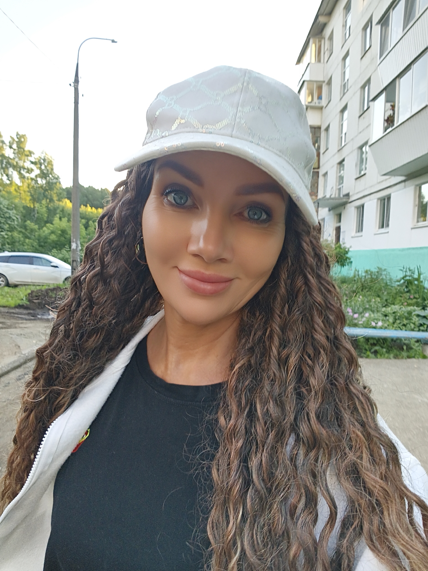 A woman with long, curly brown hair and blue eyes is taking a selfie outdoors. She is wearing a white baseball cap with a subtle patterned design and a white zippered jacket over a black t-shirt. She has on hoop earrings and her makeup appears natural. The background shows a building with white walls and multiple windows, a street lamp, some green trees and foliage, and a white car parked on the side of the road. The lighting suggests it is daytime, possibly late afternoon or early evening, with soft sunlight illuminating the scene. The woman's expression is pleasant and relaxed, and she is smiling slightly. The setting appears to be a residential area. There is no visible text in the image.