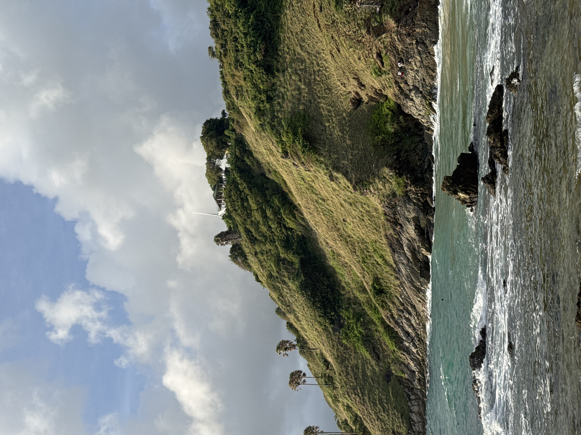 The image shows a coastal landscape with a grassy hill overlooking a rocky shoreline and the ocean. On the hilltop, a building with a white spire is visible, surrounded by trees. Several palm trees are scattered on the lower slopes of the hill. The sea is a vibrant turquoise color with white waves crashing against dark rocks in the foreground. The sky is partly cloudy, with large white clouds against a blue background. The overall scene appears to be a scenic viewpoint, possibly from Promthep Cape in Rawai, Thailand, known for its stunning sunsets and ocean vistas. There are at least three individuals visible on the rocky area of the hill.