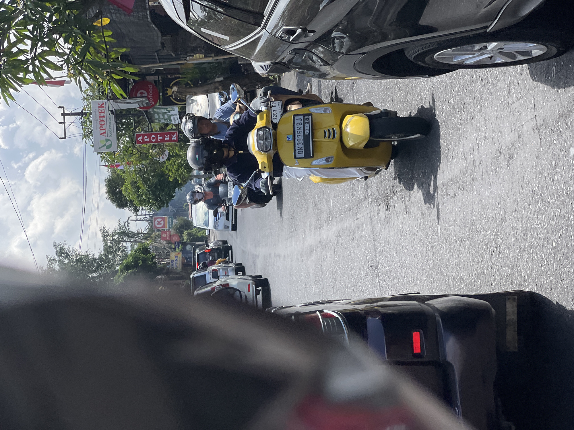 The image depicts a street scene in Ubud District, Indonesia, characterized by moderate traffic under a partly cloudy sky. In the foreground, a black SUV is partially visible on the right, with its front wheel and side panel in focus. To the left, a dark-colored vehicle, possibly a car, obstructs a significant portion of the frame, with its rear red taillight and part of its bumper visible.
The primary subject of the image is a yellow scooter, driven by a person wearing a dark helmet and a blue long-sleeved shirt, and white pants. They are wearing light-colored gloves and are looking forward. Behind them, another scooter is visible, with its rider wearing a helmet and a blue shirt. The scooters are on a paved road with visible cracks and patches, indicating wear and tear.
Several other vehicles, including cars and scooters, are in the background, forming a line of traffic. Buildings line the right side of the street, featuring signs for "APOTEK Kedewatan" and "goFood," suggesting commercial establishments. Lush green trees and foliage are present along the roadside, contributing to the tropical ambiance. The overall impression is a typical day in a bustling urban or semi-urban area of Bali, Indonesia.