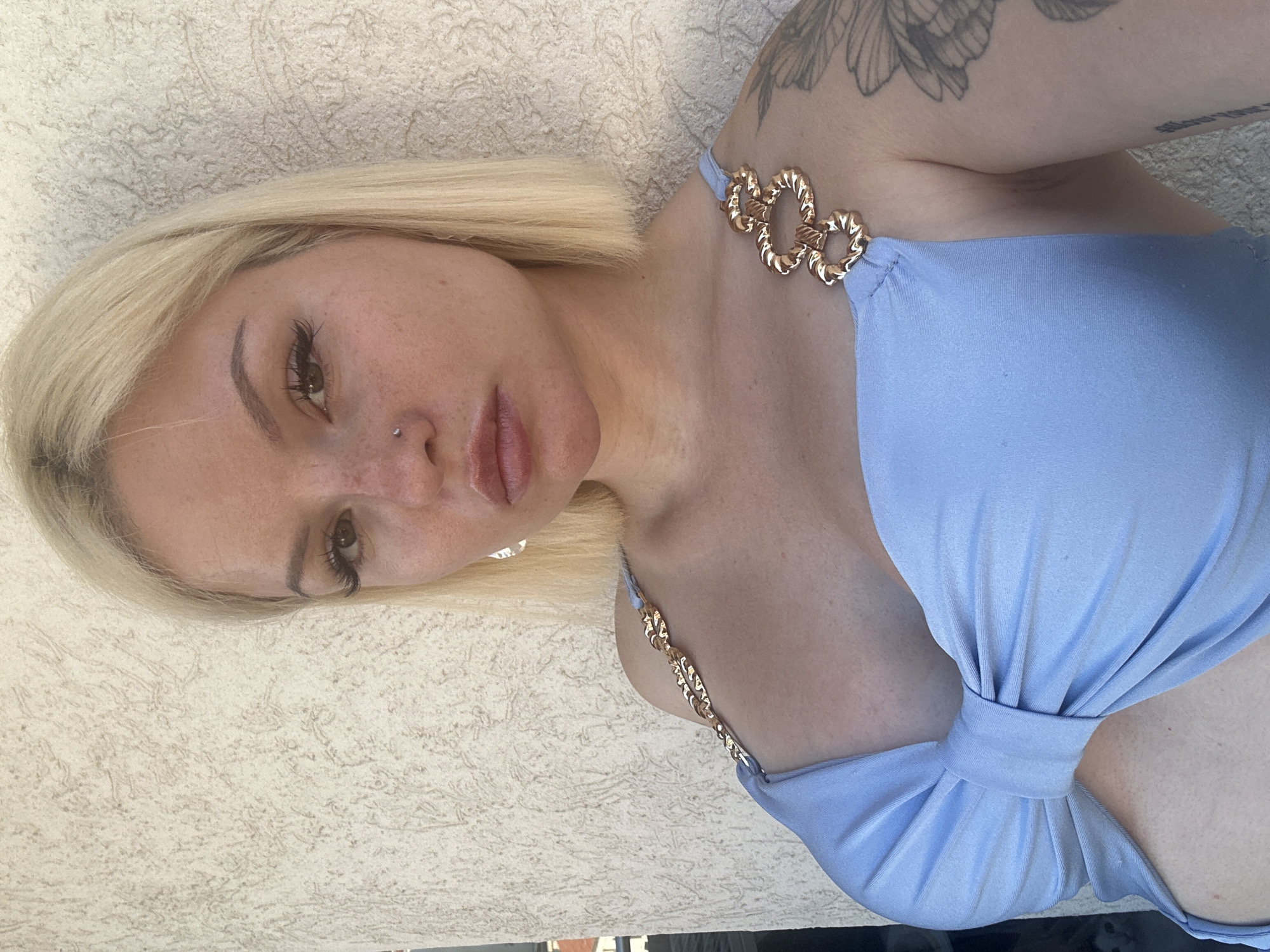 A young, fair-skinned woman with blonde bob-cut hair is captured in a close-up, sideways portrait. She is wearing a light blue bikini with gold heart-shaped accents on the straps. The woman has light brown eyes, dark eyebrows, and noticeable eyelashes. A small silver nose stud is visible, as is a tattoo on her upper left arm. Her lips are full and painted with a matte pink lipstick.
The background consists of a textured, light beige wall. The lighting suggests it is daytime, possibly outdoors, with the sun casting soft shadows. The woman's expression is neutral, with her head tilted slightly. The overall impression is a casual, sunny day portrait. There is no visible text in the image. The scene appears to be a poolside or beach setting, given the attire and outdoor lighting.