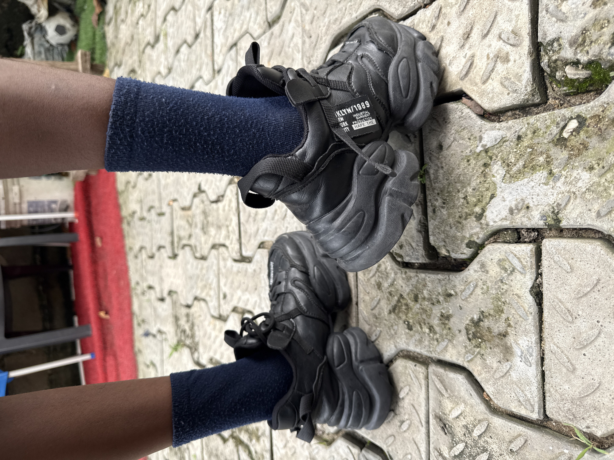 The image shows a close-up view of a person's feet wearing black chunky sneakers and dark blue socks, standing on a textured concrete pathway. The shoes have thick soles and laces, with a white tag attached to the right shoe that reads "KLYN/1989 INDUSTRIAL PRODUCTION IT MADE THE." The pathway is made of interlocking rectangular concrete blocks, some of which are stained and have moss growing in the cracks. In the background, out of focus, a red carpet is visible, along with what appears to be outdoor furniture and a soccer ball. The lighting suggests it is daytime. The scene is likely in an outdoor urban or suburban setting in Ado, Nigeria.