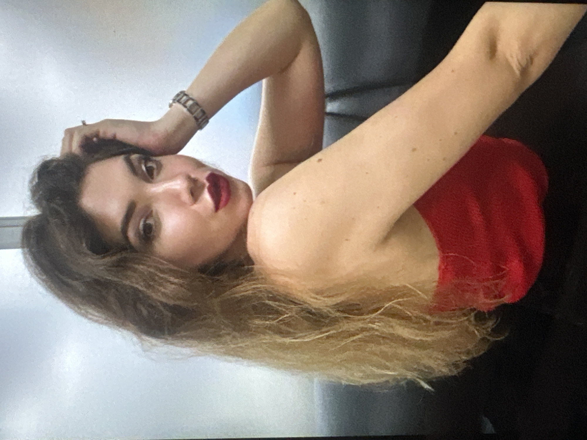 A woman with dark brown hair that fades into blonde is reclining. She is wearing a red top and a bracelet on her left wrist. She has dark red lipstick on. The location appears to be indoors against a gray background.