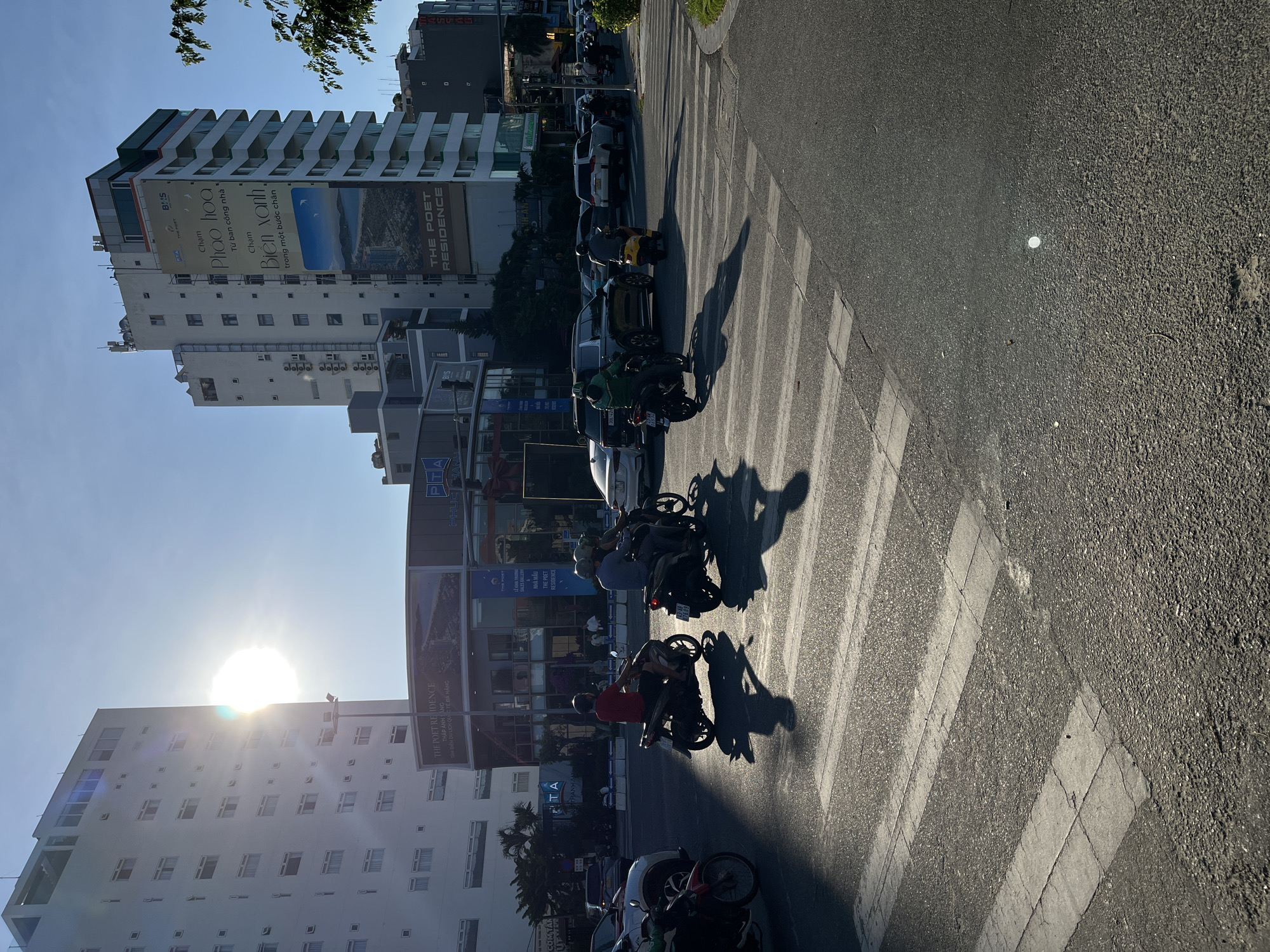 The image depicts a street scene in Đà Nẵng, Vietnam. The sun is bright in the sky. A crosswalk stretches across the road with motorcycles and scooters waiting to cross or passing through. Buildings line the street on both sides. One building is prominently marked "THE POET RESIDENCE." Another building has a large advertisement in Vietnamese, featuring images related to fireworks and the sea. Cars are parked along the side of the road. Several people are riding or waiting on the motorbikes.