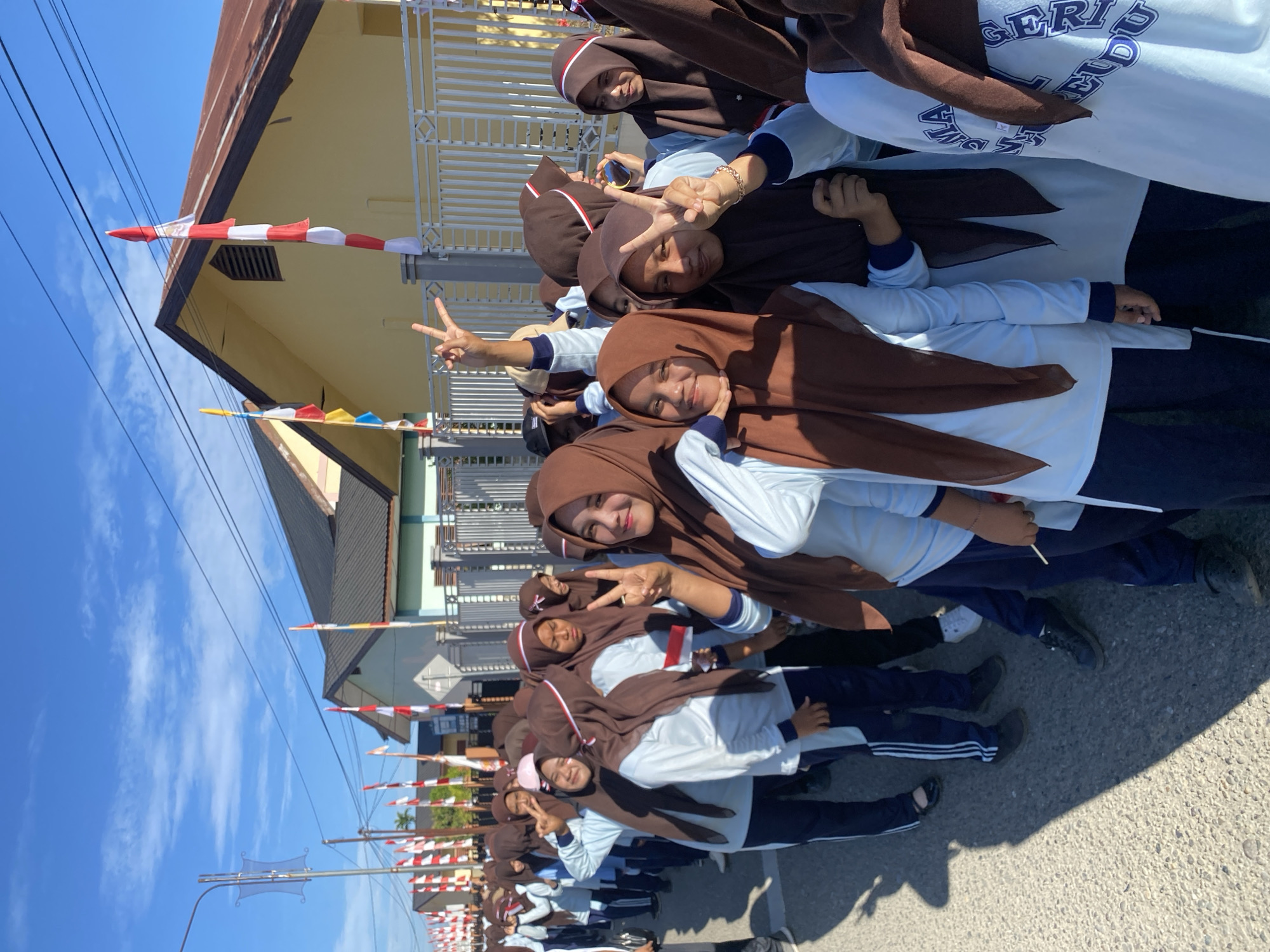 A group of young women, likely students, are lined up outdoors. They are all wearing light blue long-sleeved shirts and dark blue pants. Most of them are also wearing brown hijabs, though some have light pink or white accents on their headwear. Several of the women are smiling and making peace signs with their hands. In the background, there are houses decorated with Indonesian flags, which are red and white. The sky is clear and blue, and the sun is shining. The ground is paved with asphalt. The overall scene suggests a celebratory or patriotic event.