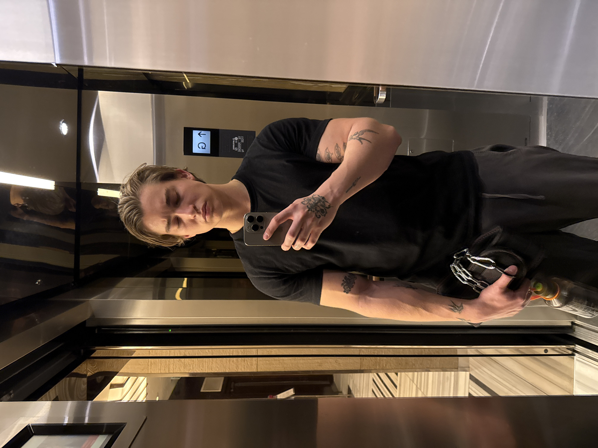 A young man is taking a mirror selfie inside an elevator. He has light-colored hair, styled back, and is wearing a black short-sleeved t-shirt and dark grey sweatpants. Numerous tattoos are visible on both of his arms, including a bird design on his left upper arm and more intricate patterns on his forearms and right upper arm.
He holds a silver-colored smartphone in his left hand, angled for the selfie. In his right hand, he holds a black hat or beanie, which has a silver chain attached, and a dark bottle with an orange cap and green label. His expression is neutral, focused on the camera.
The elevator interior is highly reflective, suggesting mirrored or polished metal surfaces. Overhead lighting fixtures are visible as reflections. To the left of the man's reflection, an elevator control panel is visible. The panel displays a downward-pointing arrow symbol and a circular arrow icon, along with some small, indistinct text or numbers below them. The lower parts of the elevator walls feature horizontal paneling, possibly wood or a similar material. The setting appears to be a well-lit, modern indoor environment.