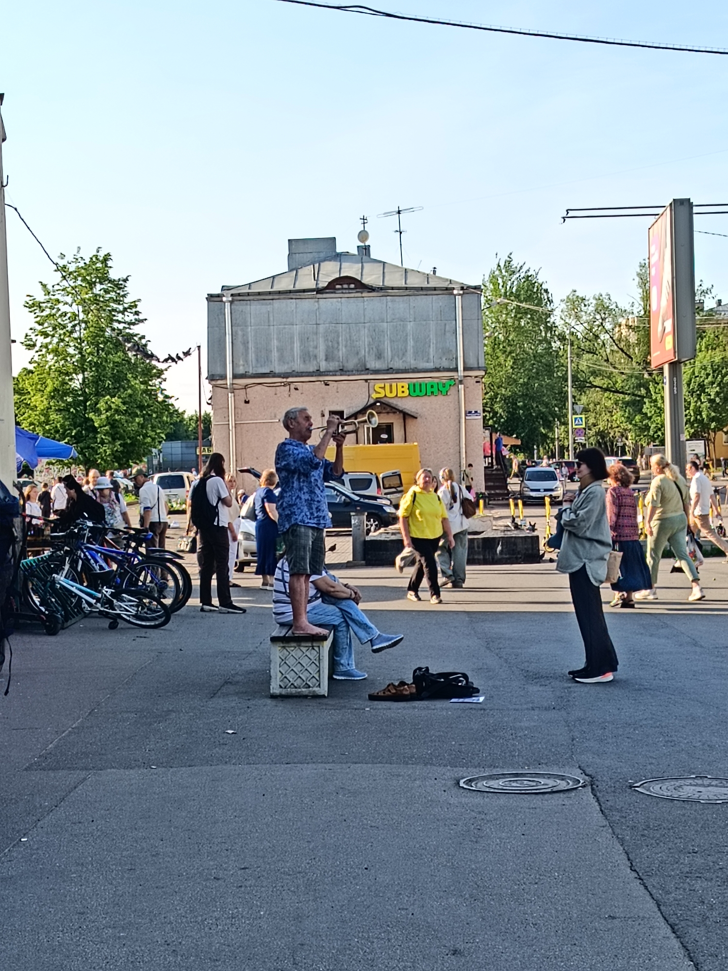 This media file depicts an outdoor scene in Saint Petersburg, Russia, likely during a market or street fair. Several people are present, engaged in various activities. In the foreground, a man stands on a decorative concrete block, playing a trumpet. He is wearing a blue patterned shirt and plaid shorts. To his left, a person is seated on the same block, wearing blue jeans and a striped shirt. A black bag and sandals are on the ground nearby.
Further back, a crowd of people is milling about. Some are walking, others appear to be browsing or socializing. Bicycles are parked to the left of the frame. Behind the crowd, a building with a "SUBWAY" sign is visible, suggesting an urban environment. The lighting indicates it is daytime, likely late afternoon or early evening, with the sun casting long shadows. The weather appears pleasant. The overall atmosphere seems relaxed and social.
Notable details include the architecture of the buildings, the presence of bicycles, and the "SUBWAY" sign which helps contextualize the location as an urban area in Russia. The people are dressed in casual summer clothing. The activity appears to be a public gathering, possibly a street market or festival, with a musician entertaining the crowd.