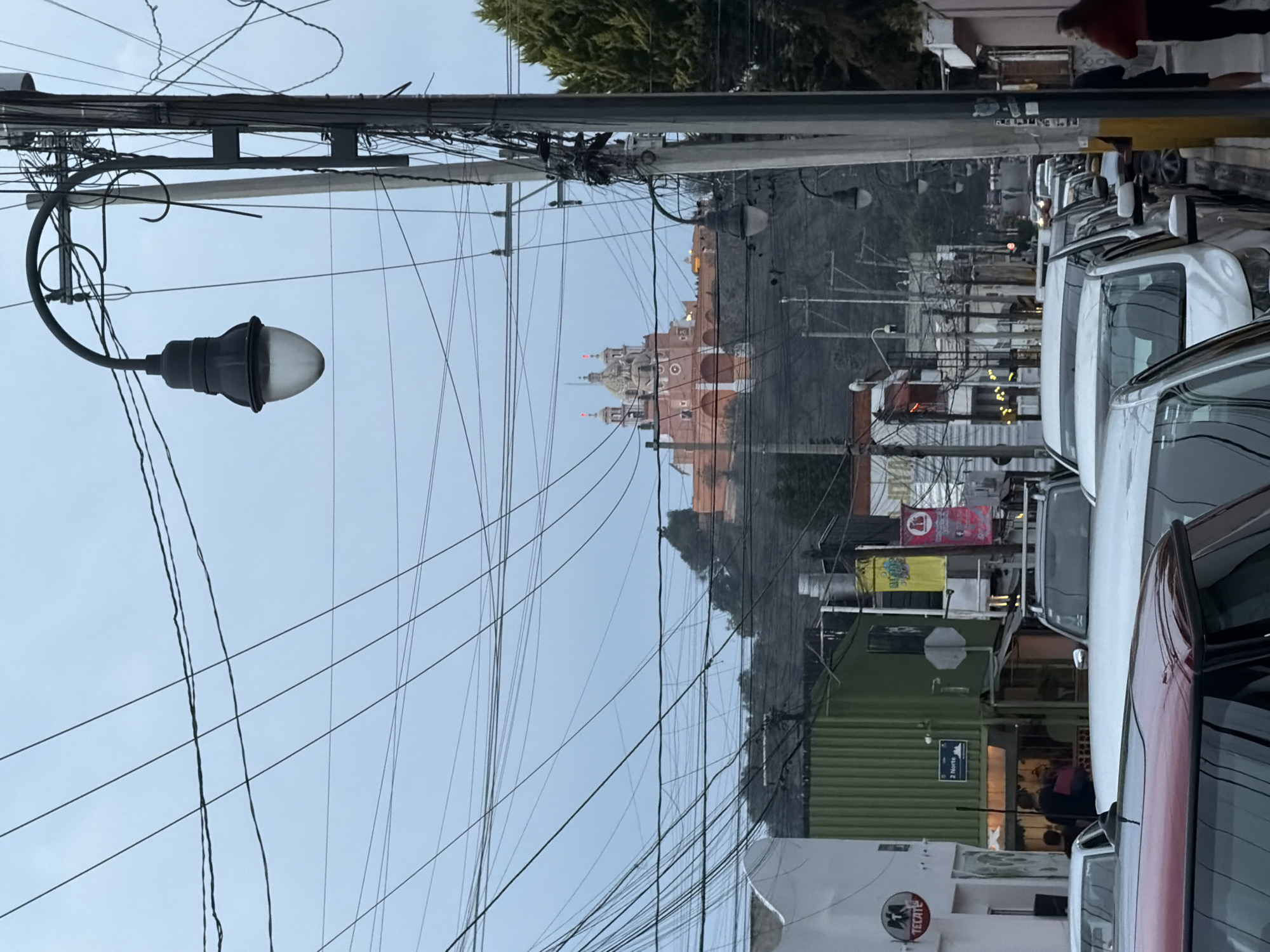 The image depicts a street view in San Andrés Cholula, Mexico, characterized by a dense overhead network of electrical and communication wires. On the left side of the frame is a street lamp with a rounded glass globe, attached to a utility pole. A multi-domed, red-colored building is visible in the distance, situated on a slight hill, likely a church or religious structure. The right side of the image shows a row of parked cars, with pedestrians passing by on the sidewalk. Commercial establishments are visible between the cars and utility poles, identifiable by signs and building architecture. The sky is overcast, suggesting an early morning or late afternoon scene.