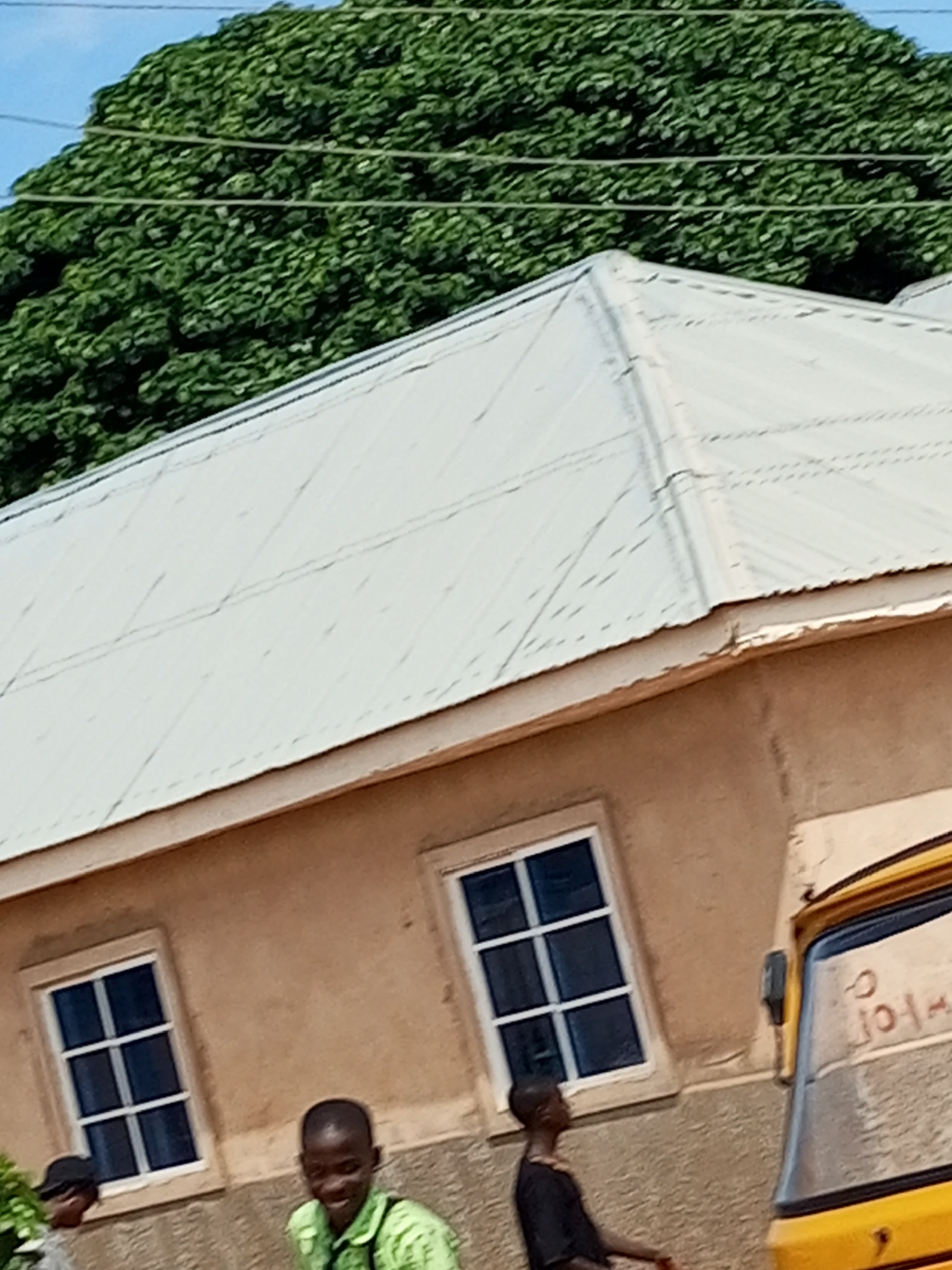 In Tempure, Nigeria, a weathered, light-colored building with a corrugated metal roof is observed. Two dark-framed windows with blue-tinted glass are visible on the building's tan facade. In front of the building, three individuals are present. One young person, wearing a lime green shirt, is smiling and looking towards the camera. Another individual, wearing a black t-shirt, is standing to the left of the first person and looking away. A third person, wearing a black hat, is partially visible on the far left. A yellow auto-rickshaw is parked to the right of the building, with red markings on its windshield. Lush green trees form a dense canopy at the top of the image, with thin wires running across them. The ground in front of the building appears to be a dusty, uneven surface.