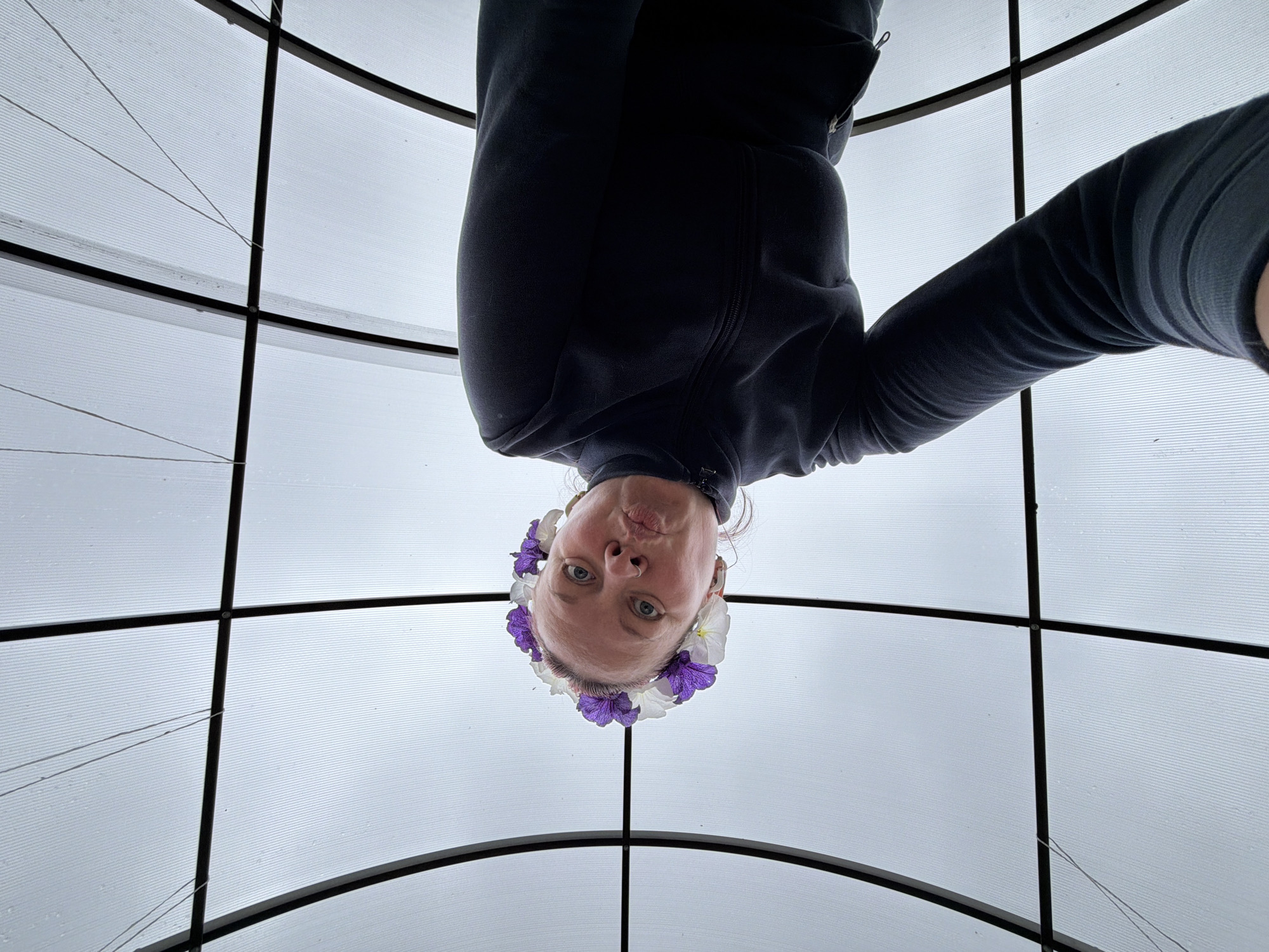 A low-angle shot depicts a person wearing a dark-colored zip-up jacket and a flower crown with purple and white flowers. The person is upside down in the frame. The background is a gray-white structure with a grid pattern of black beams. The location is in the city of Chaikovsky, Russia.