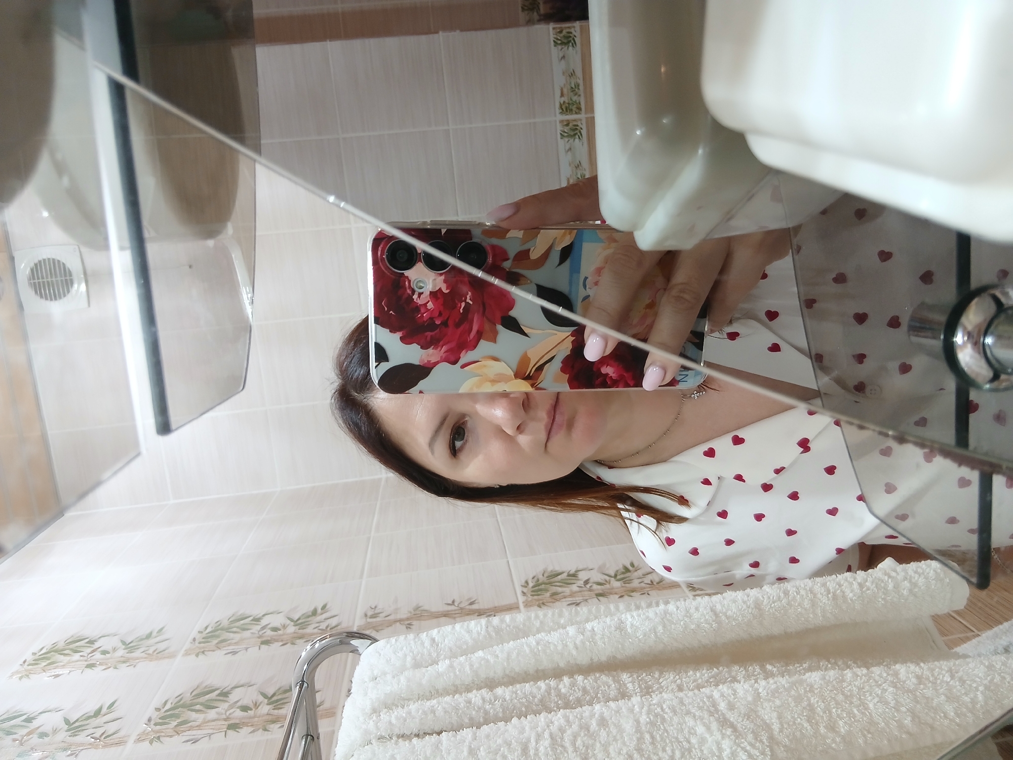 A woman is taking a mirror selfie in a bathroom. She has dark hair and is wearing a white pajama top with red hearts. Her phone, with a floral case, is held up to capture the reflection. To her left, a white fluffy towel hangs on a rack. The bathroom walls are tiled with a bamboo pattern. A vent is visible on the ceiling.