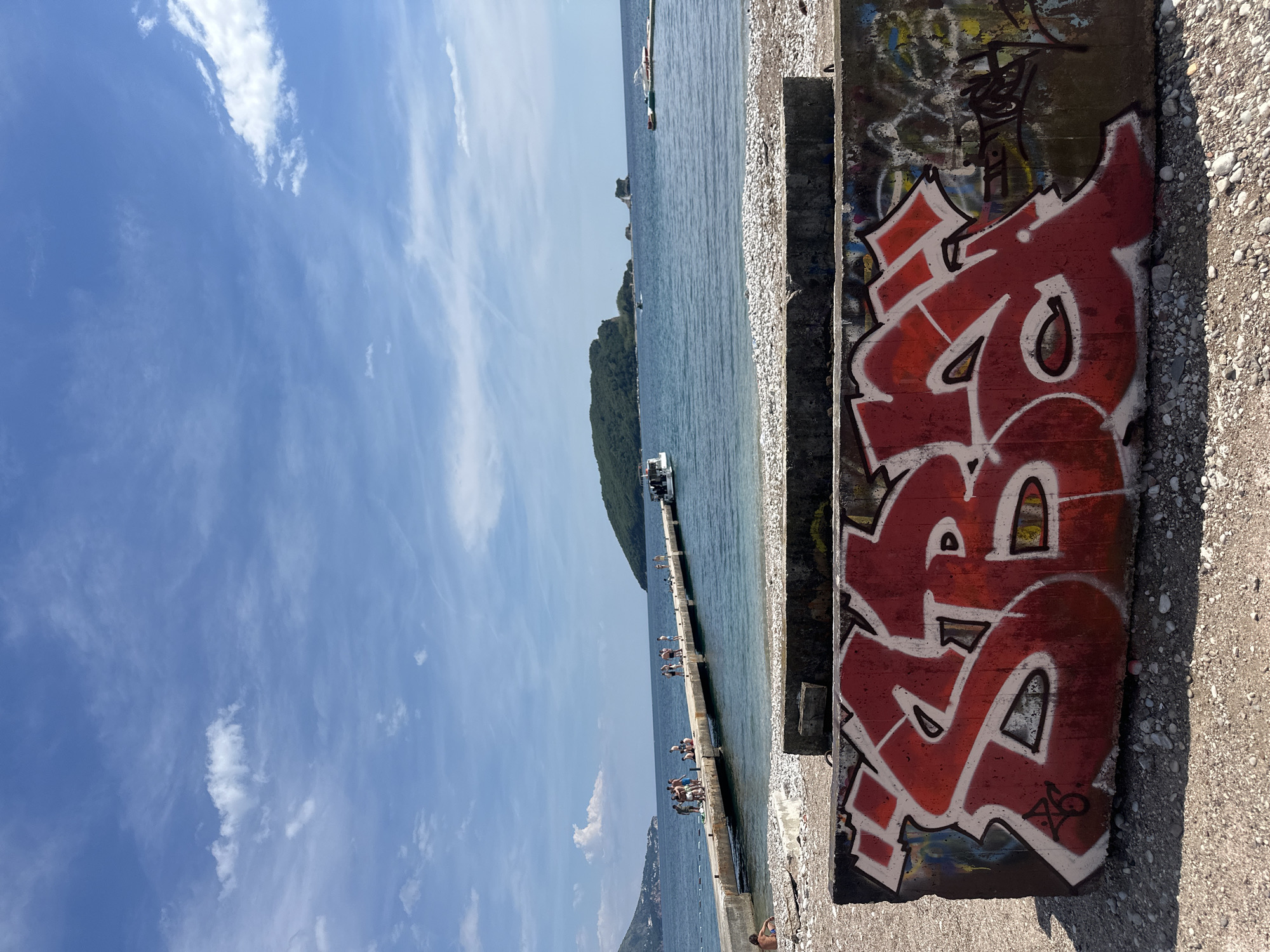 A sideways shot shows a concrete pier extending into the Adriatic Sea, with a rocky beach and graffiti-covered concrete structures on the right, located in Budva, Montenegro. The sky is blue with scattered clouds. Several people are on the pier, some standing and others on boats at the end. The graffiti on the concrete barriers is predominantly red with white outlines, and some additional black graffiti can also be seen. The scene gives a view of a coastline and an island covered in vegetation.