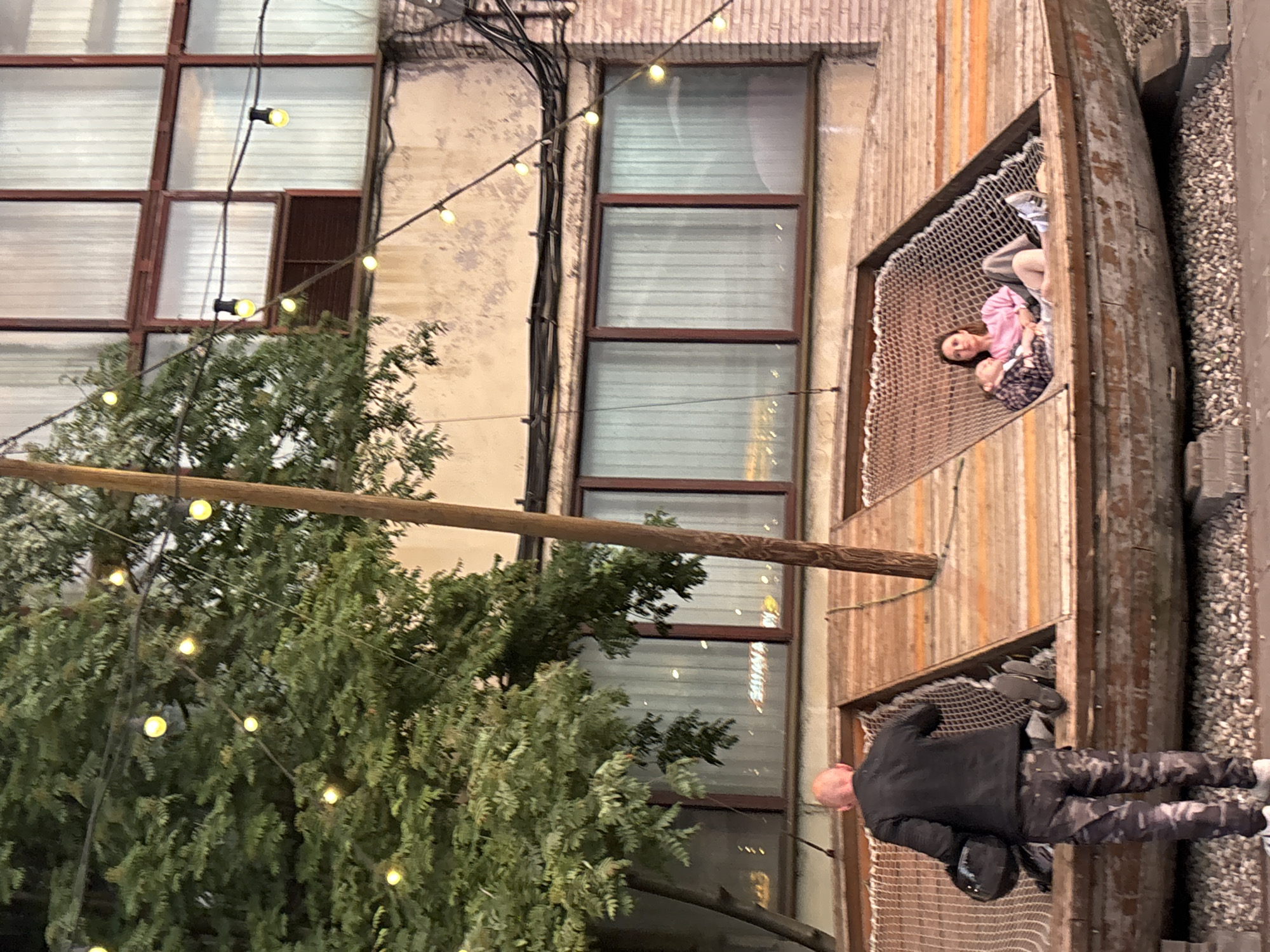The image depicts an outdoor seating area in Saint Petersburg, Russia. In the foreground, a bald man in a black jacket and camouflage pants is walking away from the camera, holding a black motorcycle helmet. He is approaching a wooden structure resembling an upside-down boat, where two people are seated in a net hammock. A woman in a pink sweatshirt is on the right, and a child is lying down next to her. The wooden structure is elevated and appears to be a unique architectural feature.
Behind the seating area, a building with large windows covered by sheer white curtains is visible. String lights with yellow bulbs are draped across the scene, adding a festive atmosphere. Lush green trees with dense foliage frame the left side of the image, and a wooden pole extends vertically through the center. The ground around the structure is covered in small grey pebbles. The overall impression is a relaxed, urban outdoor space with an artistic touch.