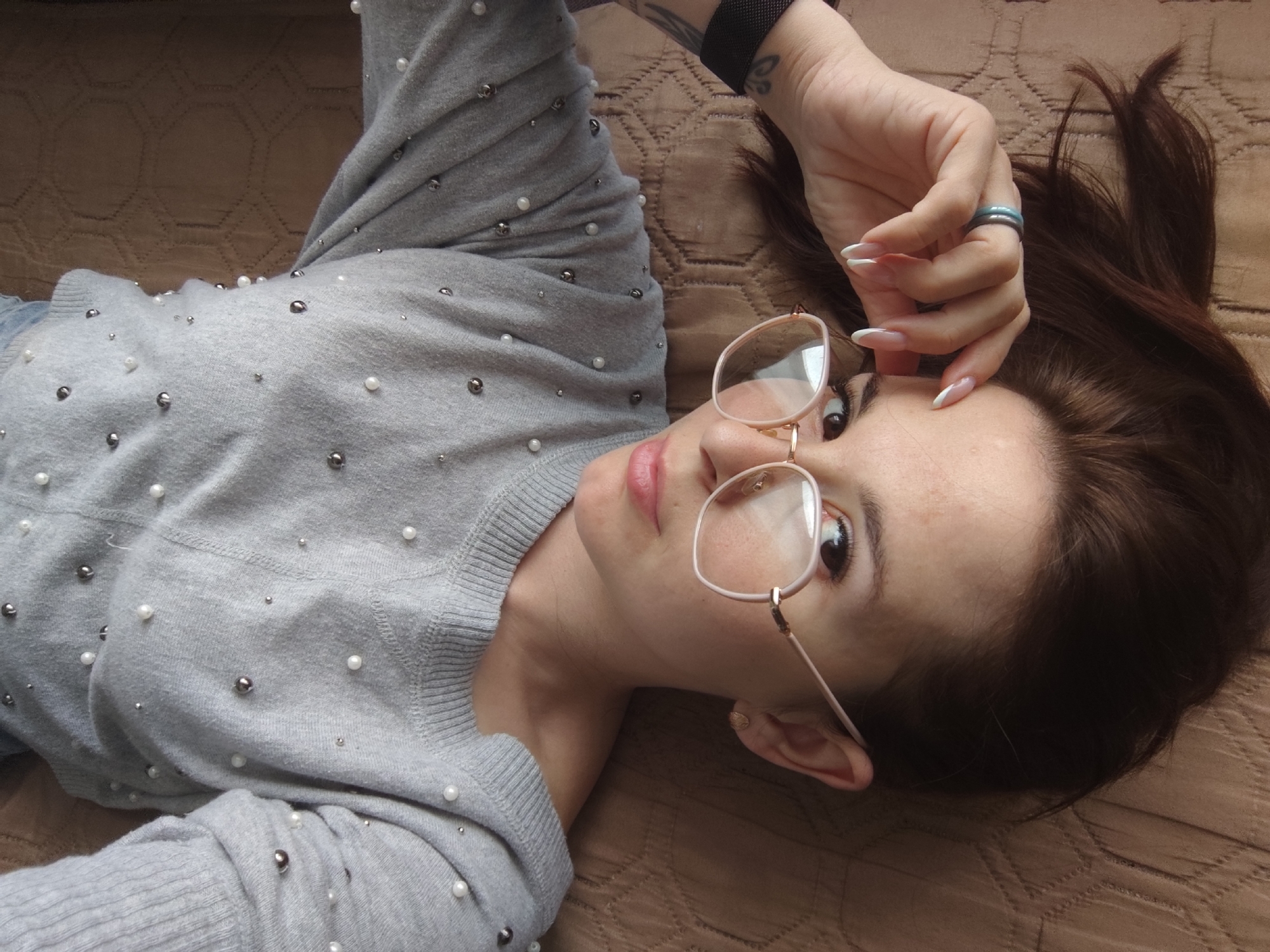 A person is depicted from an overhead angle, lying on a textured brown surface, possibly a bed or couch. The individual has light brown hair, which is fanned out behind their head, and is wearing thin-rimmed glasses. Their eyes are open, looking slightly upwards towards the viewer.
The person is dressed in a light grey sweater adorned with an array of small white pearls and metallic silver beads. Their left hand is raised towards their face, with fingers gently touching the side of the glasses. The fingernails are long and have a classic French manicure. A light blue ring is visible on the ring finger of the left hand, and a dark, possibly black, smartwatch or fitness tracker is worn on the left wrist. A small, dark tattoo can be seen on the inner part of the left forearm.
The setting appears to be indoors, illuminated by soft, diffused light, suggesting daytime. The background surface features a repeating geometric pattern, adding texture to the scene. There are no overt geographical cues visible in the image itself, though the provided context indicates "Фоки, Russia." No visible text is present in the media file. The overall mood is calm, with the individual presenting a neutral or slightly contemplative expression.