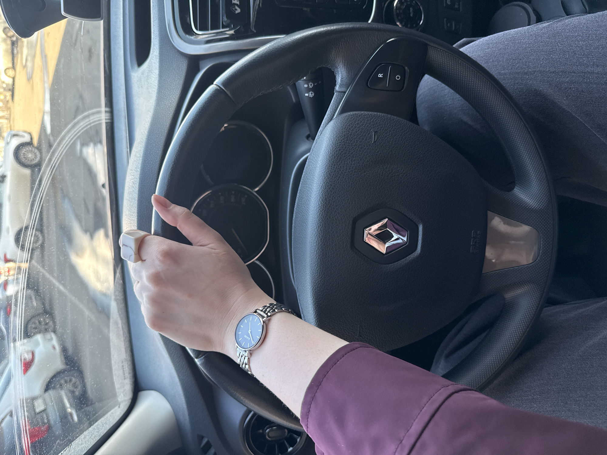 A person is driving a Renault car, possibly in a parking lot in the city of Izhevsk, Russia based on the blurred background. The person is wearing a maroon shirt, a silver watch, and a white ring on their left hand. The car's steering wheel is visible with the Renault logo. The dashboard and windshield of the car are also visible.