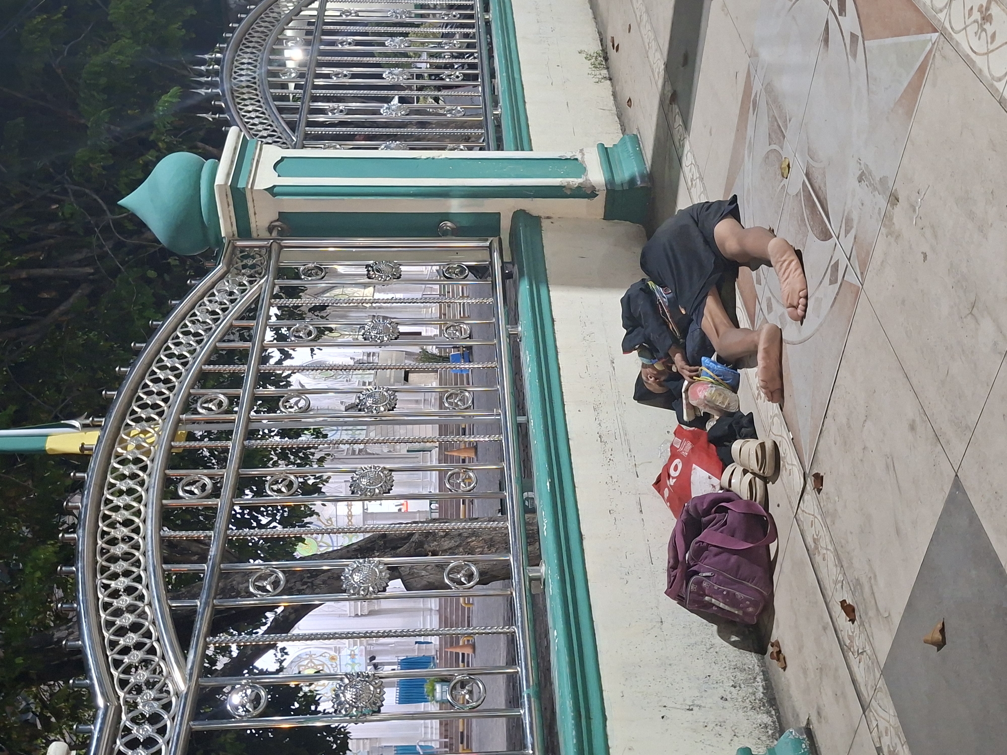 A person is prostrated in prayer on a tiled surface next to a metal and green concrete fence. Personal belongings, including a purple bag, red bag, and sandals, are beside the person. The fence, with decorative metal elements, lines a building with blue accents. The scene appears to be in Lhokseumawe, Indonesia, based on architectural styles. The photograph is taken during the day or dusk, with a mix of artificial light and natural light.